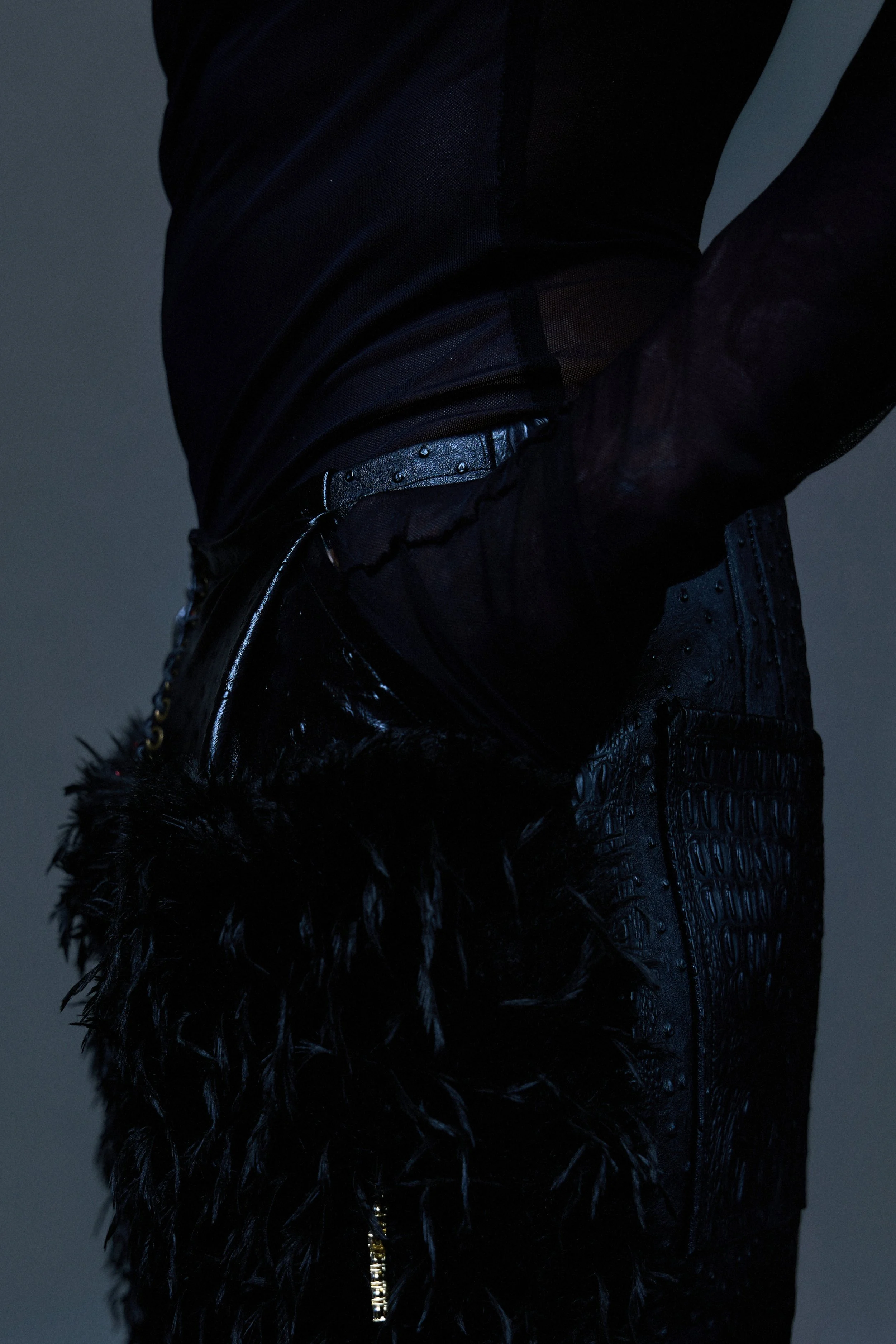 Close-up of a person's side, wearing a black, shiny, textured jacket with feathered details on the sleeves, a matching black top, black pants, and a shiny black belt with metal studs.