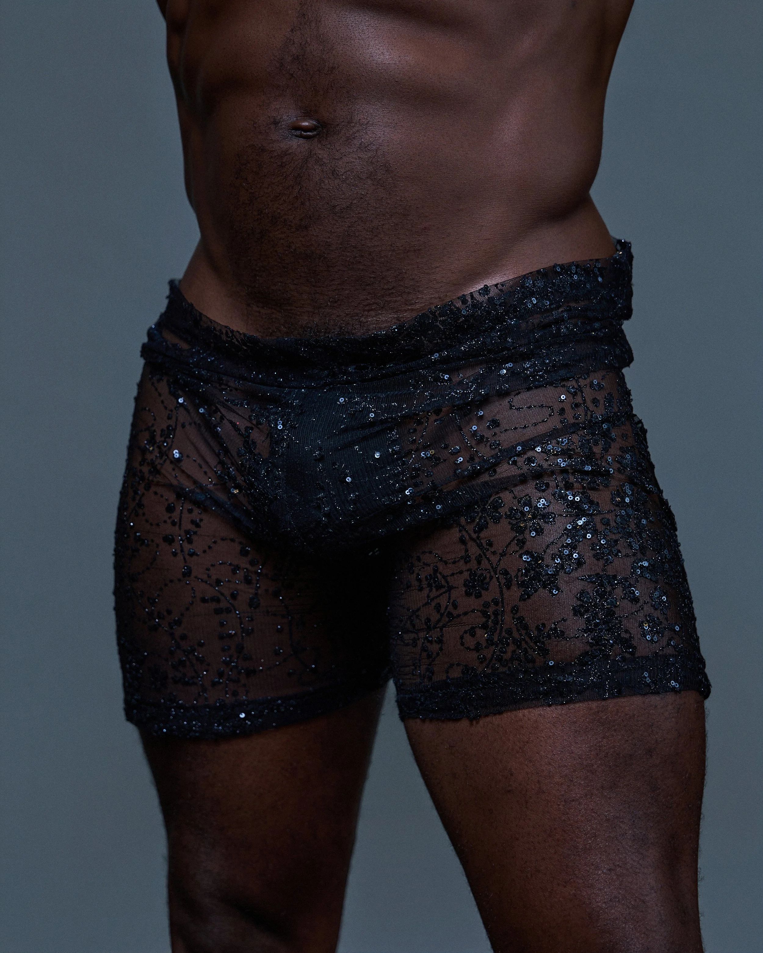 Close-up of a dark-skinned person's midsection wearing black sequin shorts, with a focus on the stomach and a hint of pubic hair.