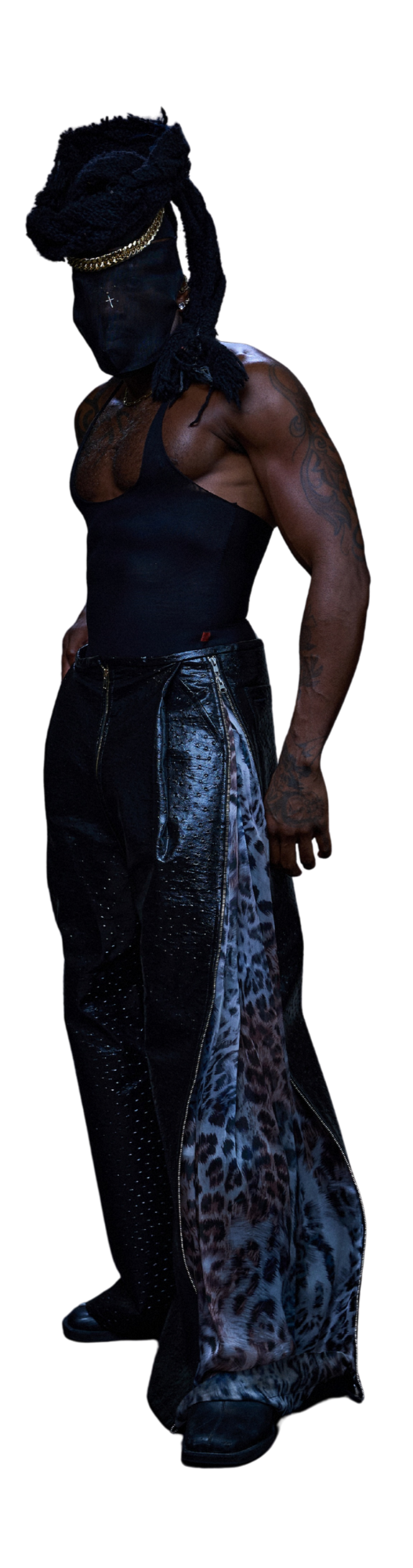 A person with tattoos wearing a black tank top, leopard print pants with a side zipper, a black mask with white markings, dreadlocks, a gold chain, and standing against a dark background.
