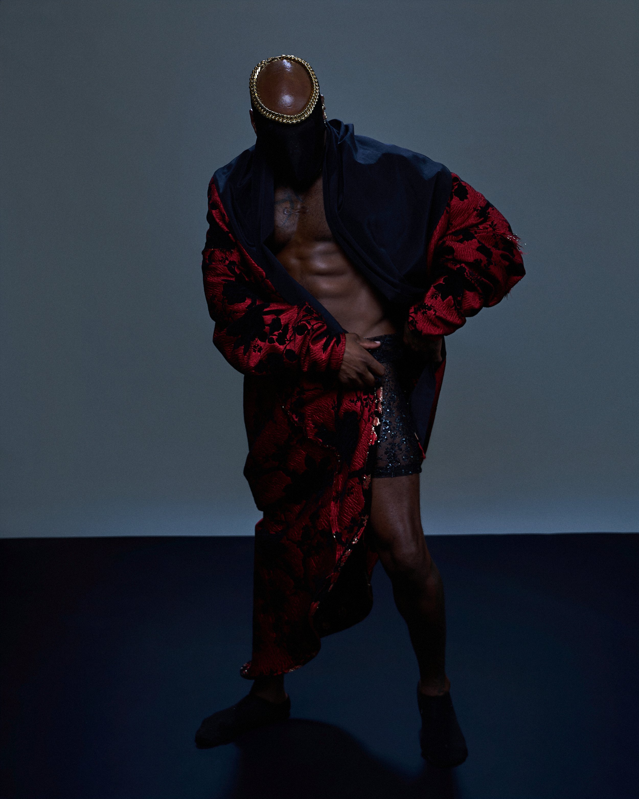 A shirtless man with a dark skin tone is wearing a black jacket with red and black floral pattern, and a gold chain headband. He is standing against a plain dark background with one hand on his waist and is wearing black shoes.