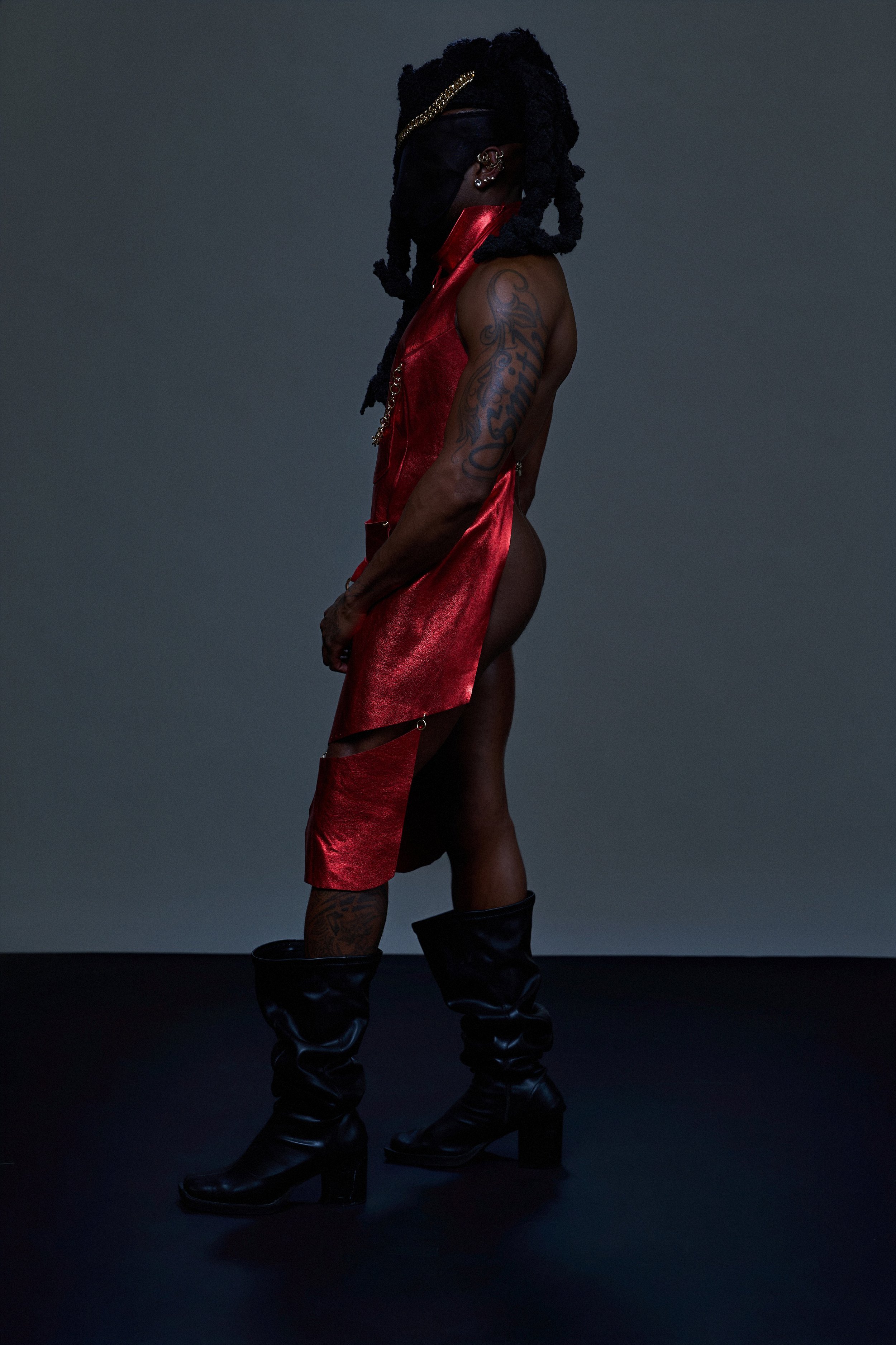 Person with dreadlocks wearing a black mask, red satin dress, and black boots, standing against a dark background.