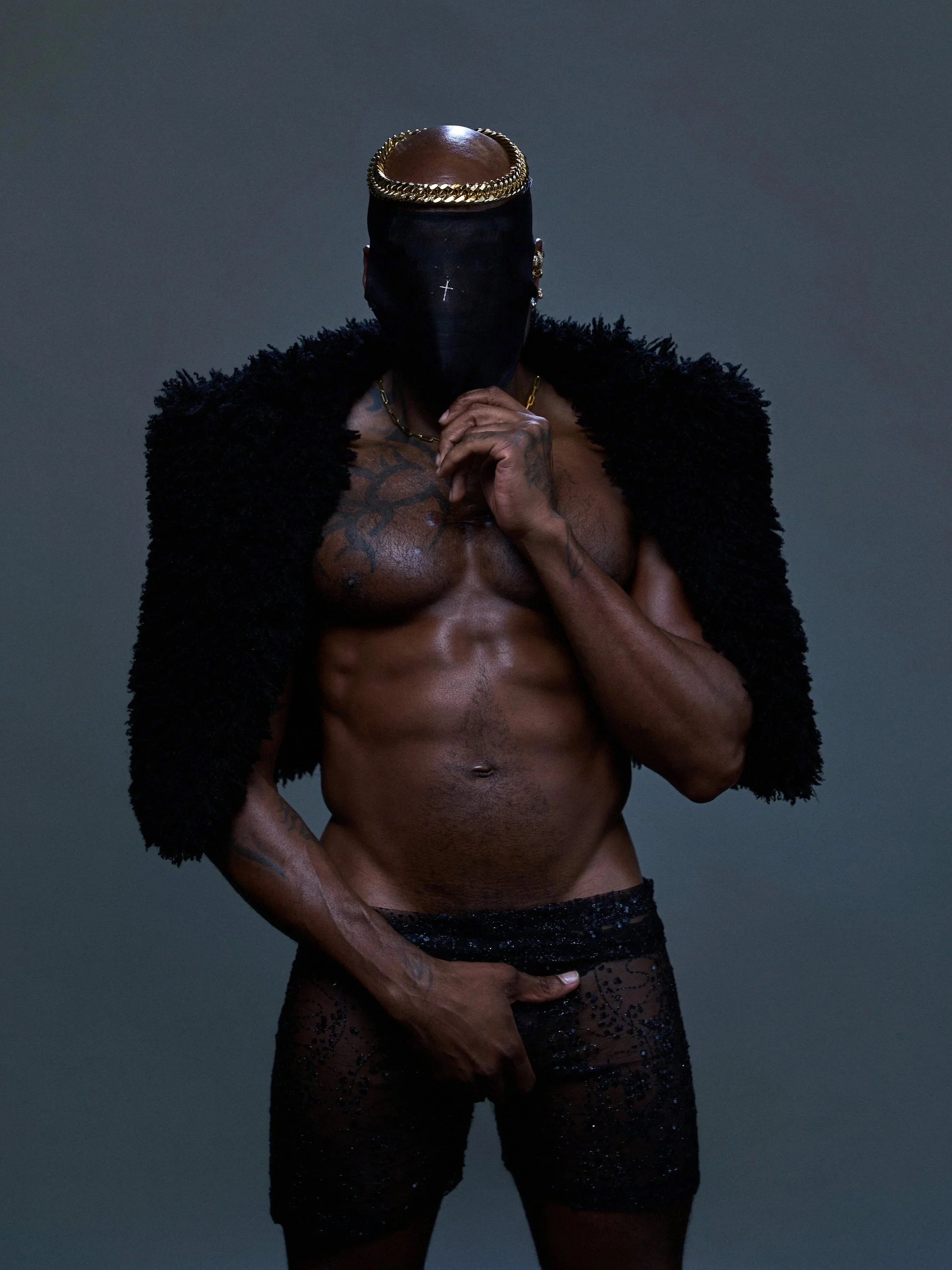 A shirtless man with tattooed torso and arms, wearing black lace underwear, black furry jacket, gold chain, and gold hoop earrings, with his face covered by a black mask and a gold chain headpiece, standing against a dark grey background.