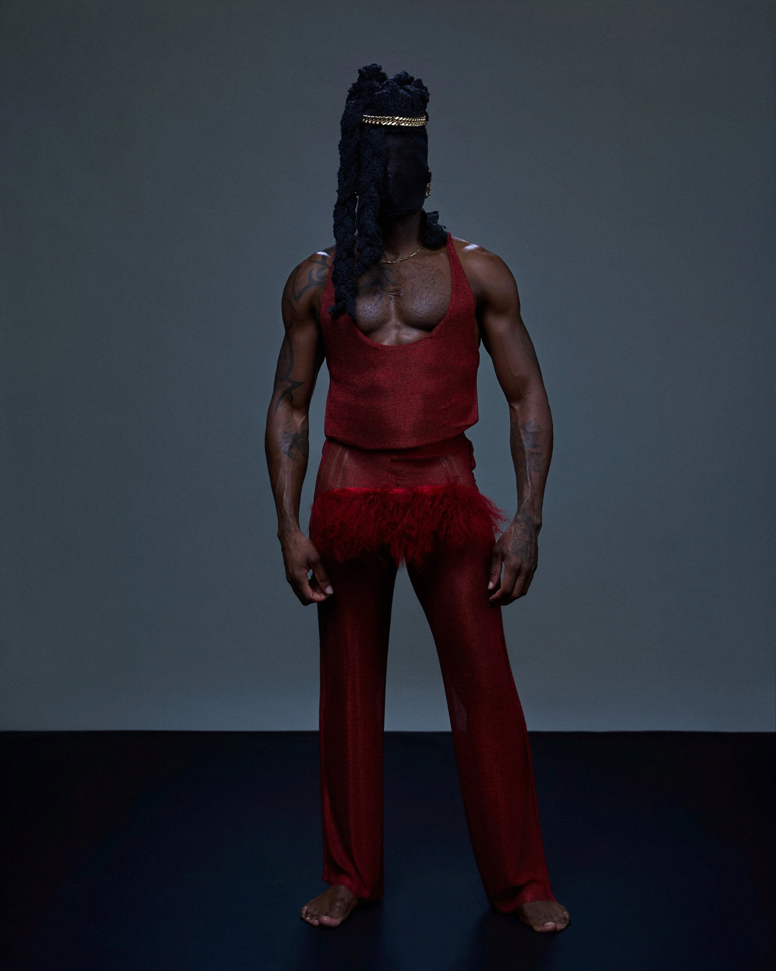 A person with dark skin and long dreadlocks, wearing a red sleeveless top and matching red pants with sheer fabric at the bottom, standing barefoot against a plain dark background. The person's face is obscured by a black cloth.