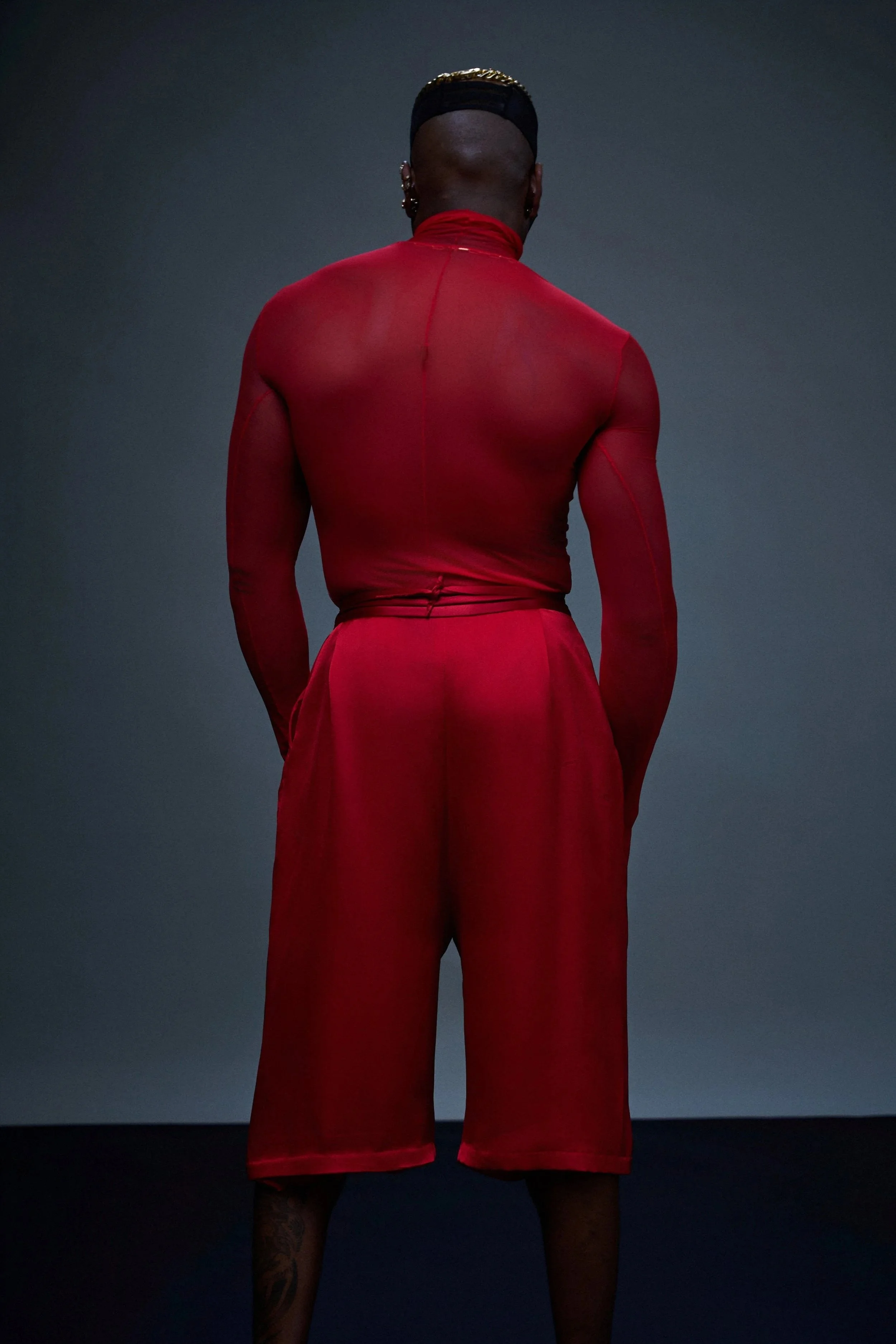 Back view of a person with a shaved head, gold chain on their head, multiple earrings, wearing a red sheer long-sleeve top and red pants, standing against a dark background.