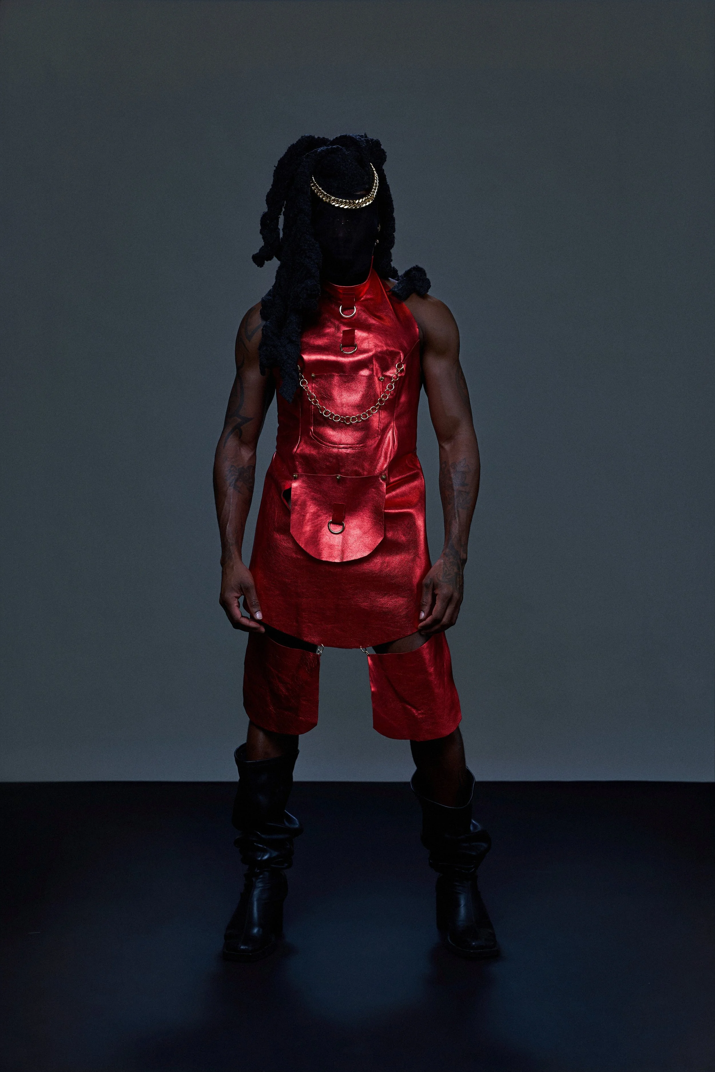A person wearing a red leather dress and pants, black boots, and accessories, with a black face covering, standing against a plain gray background.