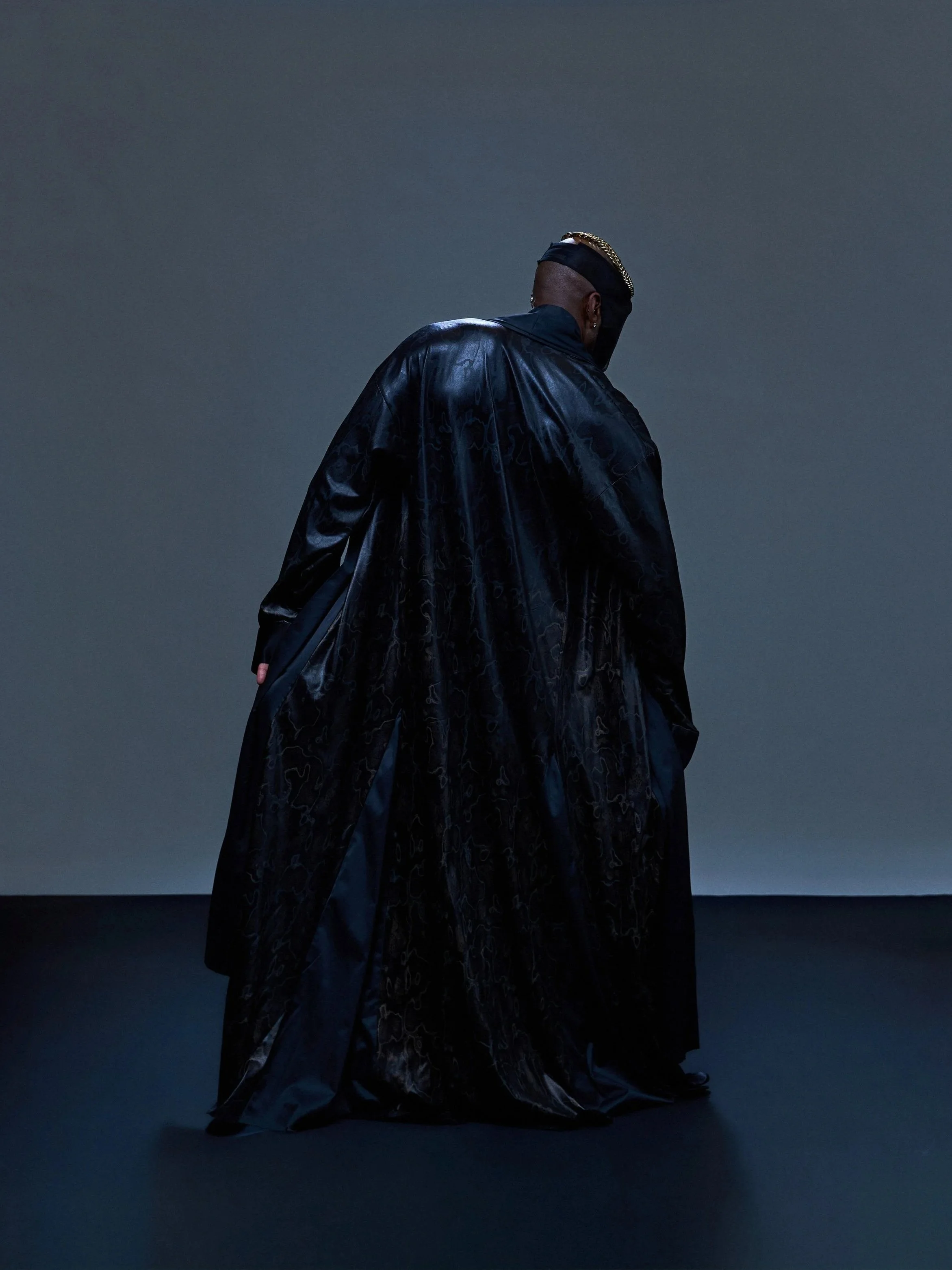A person with a mohawk hairstyle, wearing a shiny black coat, standing on a dark stage with a gray background, facing away from the camera.