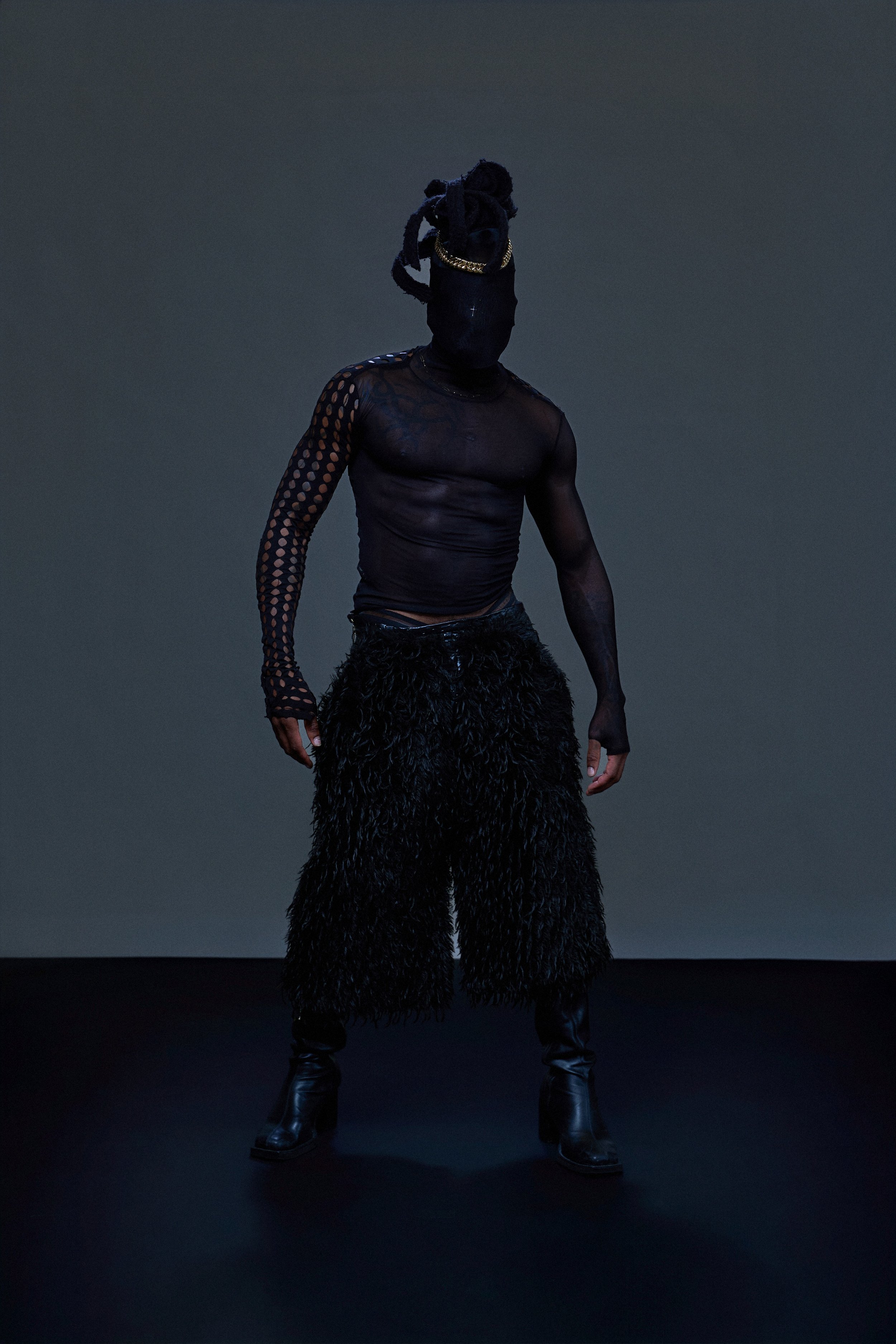 Geoffrey Smith - Model with black mask covering face, gold chain, black mesh top with polka dot sleeves, furry pants, and black boots standing on a black floor against a dark background.
