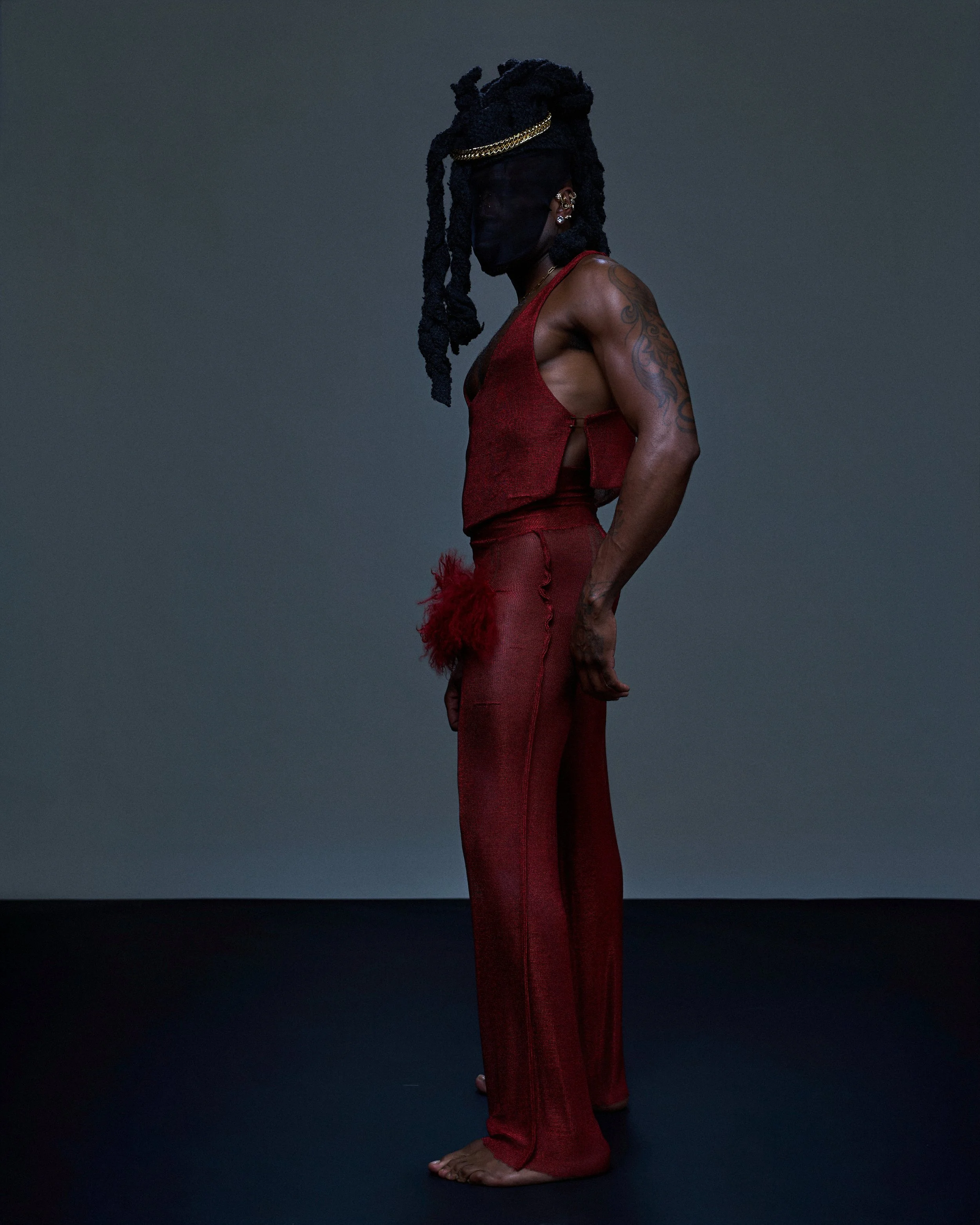Woman with dark skin and tattoos wearing a red sleeveless top and matching pants, barefoot, standing sideways against a plain dark background, with her face obscured by a black face mask, her hair styled in braided dreadlocks with some tied up.