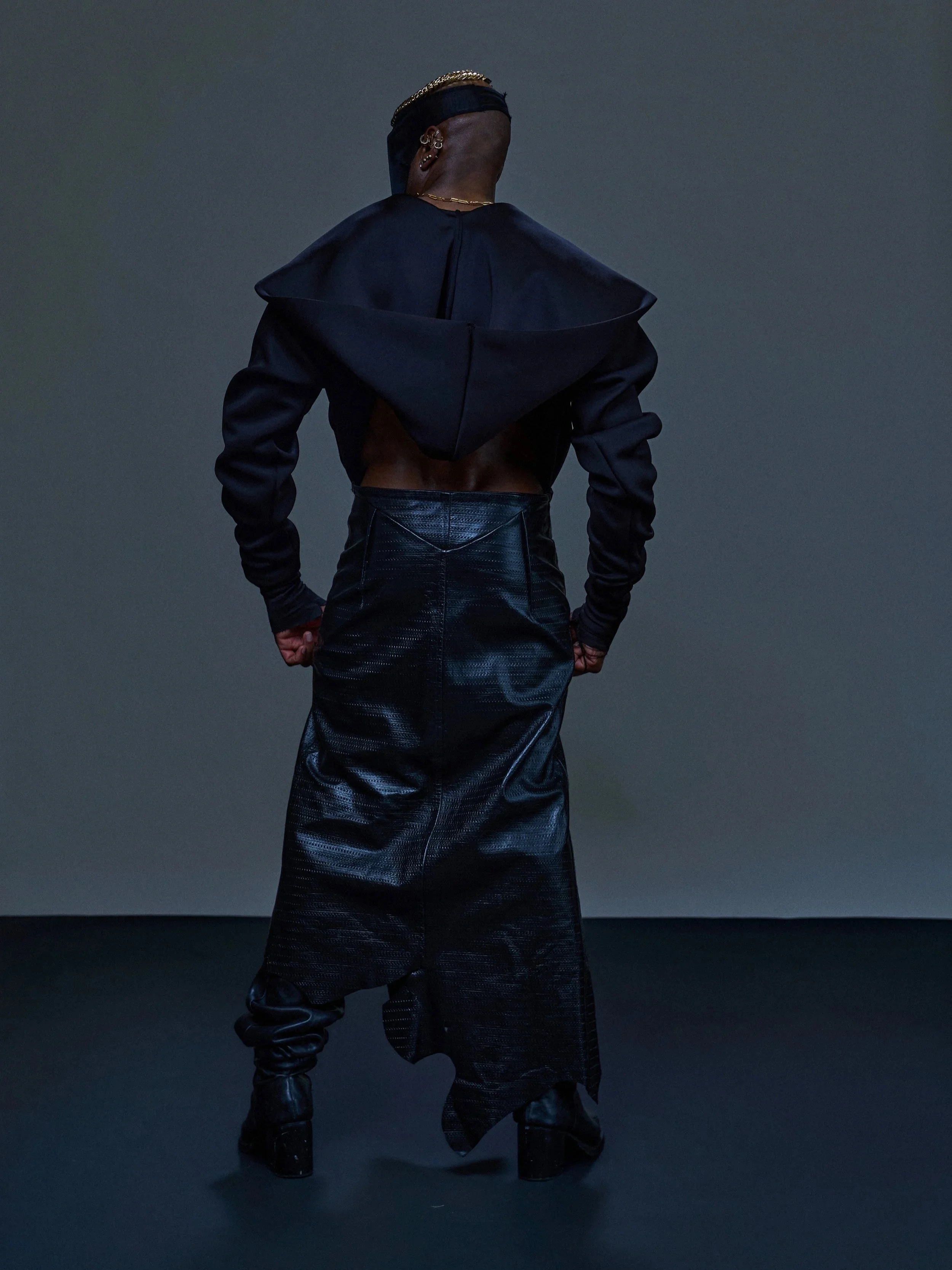 Person wearing a black hooded jacket with large lapels, shiny black pants, and black boots, facing away against a dark background.