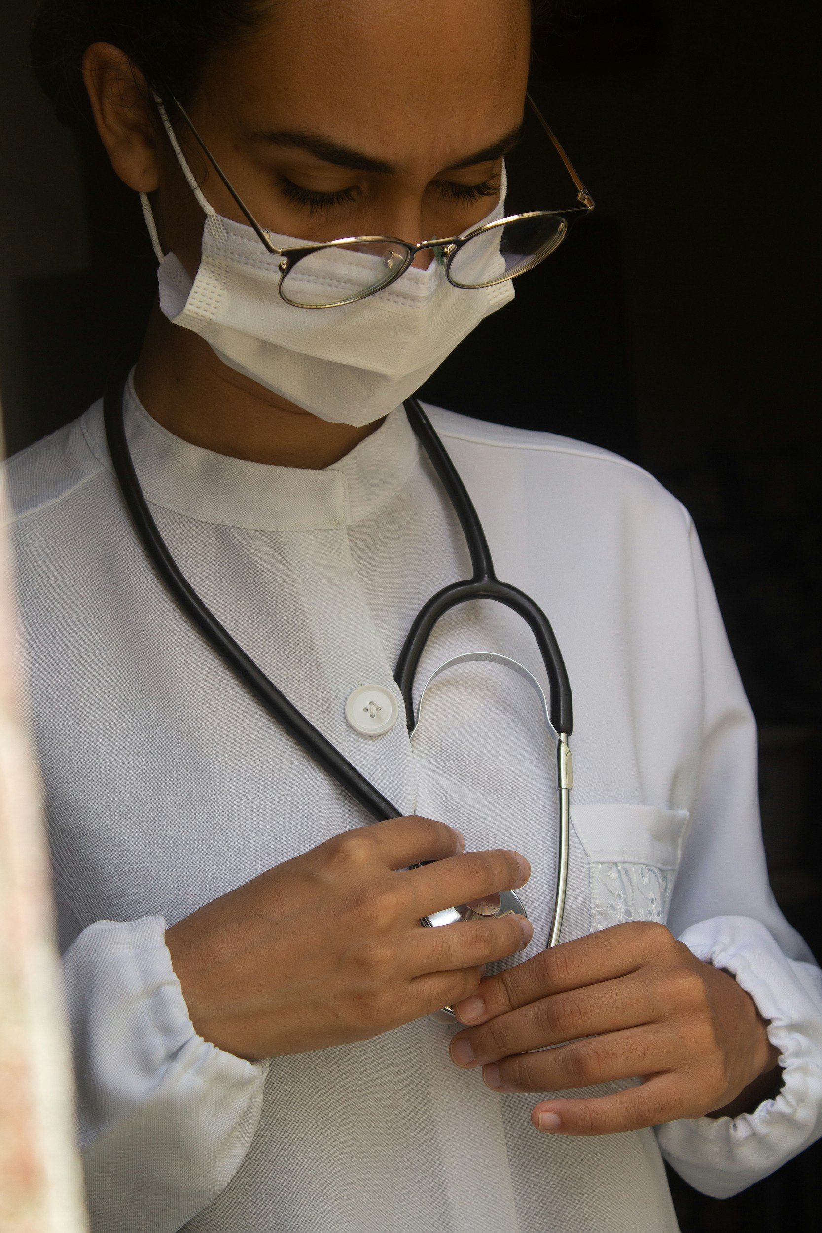 A healthcare professional, wearing glasses and a face mask, is holding a stethoscope on their chest with both hands, wearing a white uniform.