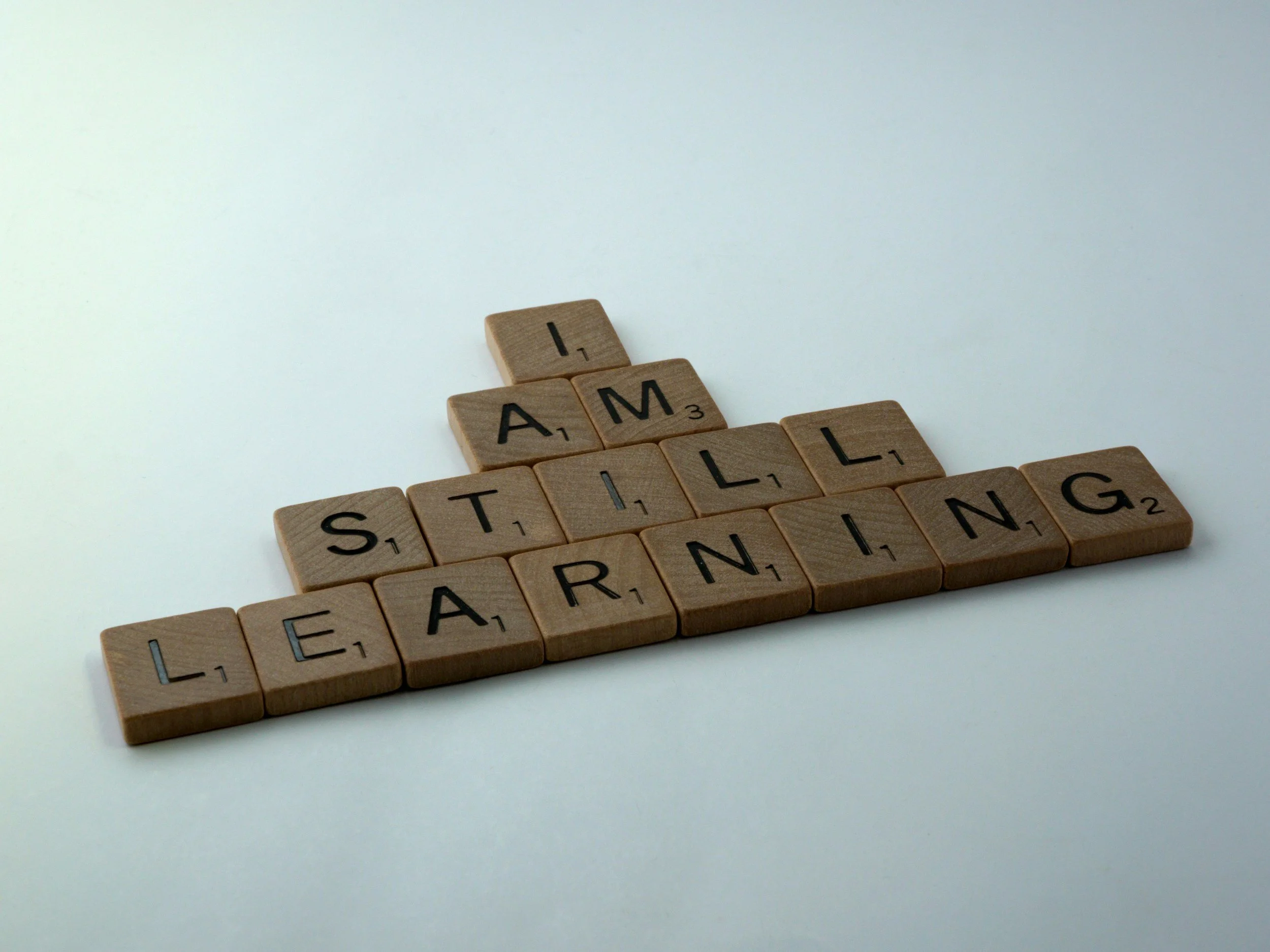 Wooden Scrabble tiles spelling the words 'I AM' and 'STILL LEARNING' on a white background.