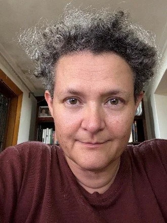 A person with curly hair and light skin taking a selfie indoors, wearing a maroon shirt, with bookshelves in the background.