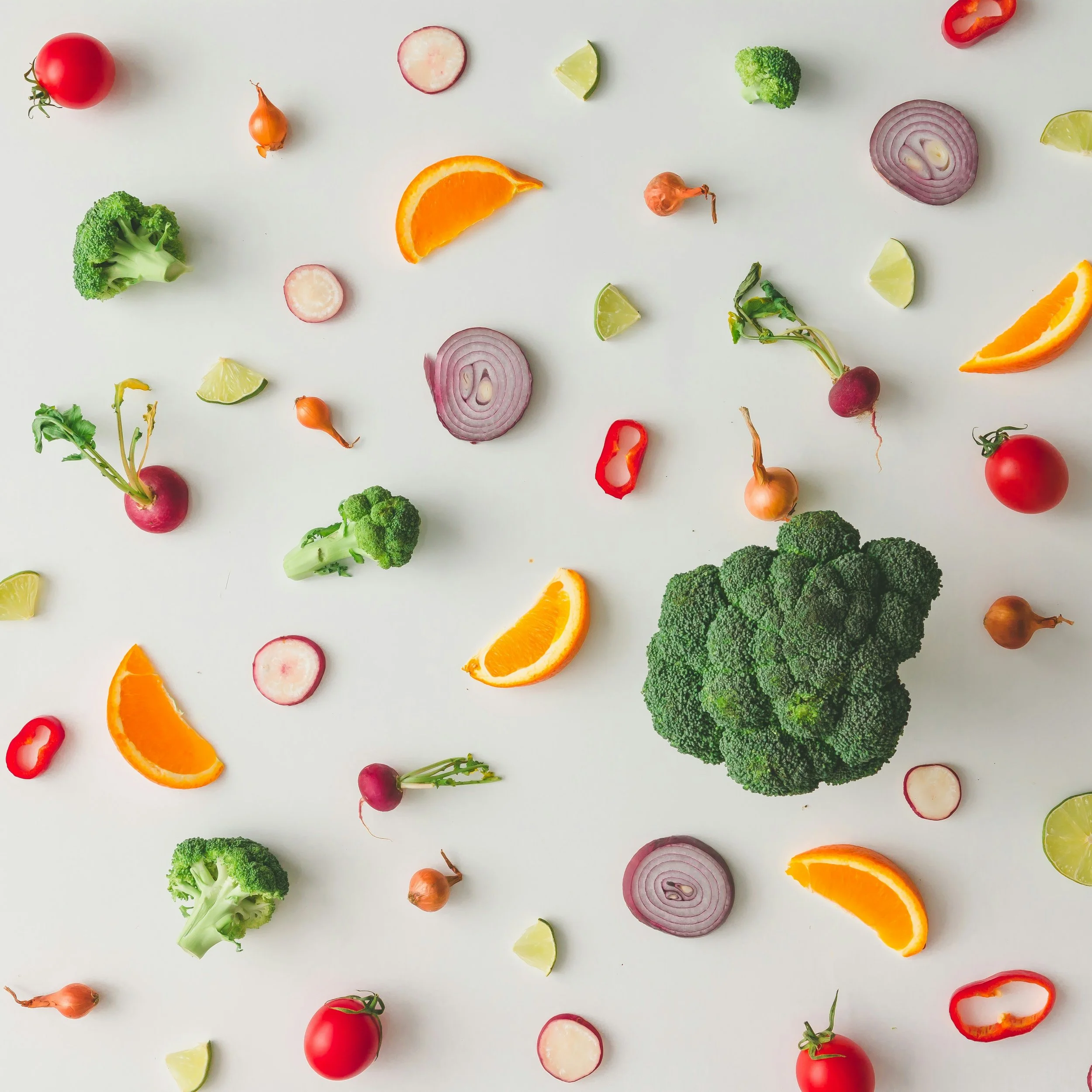Assorted vegetables and citrus slices on a white background, including tomatoes, broccoli, radishes, red onion, lime wedges, lemon wedges, and orange slices.