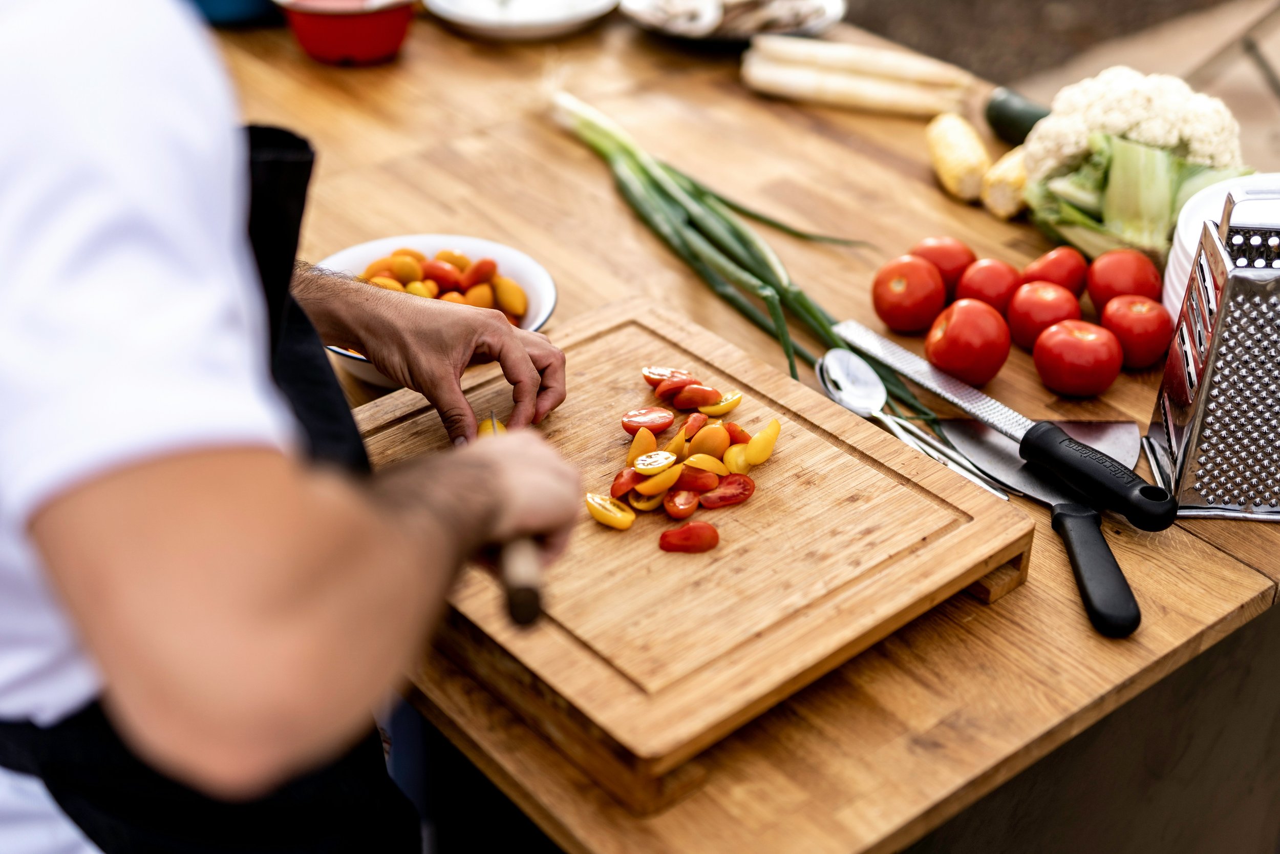 A chef is preparing cherry tomatoes on a wooden cutting board surrounded by fresh vegetables supplied by Farmers Bounty including cauliflower, garlic, and zucchini grown on their farm in Werribee. kitchen.