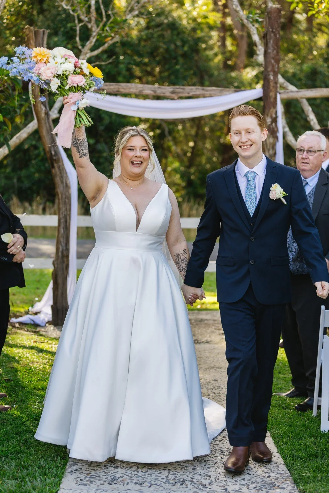A bride and groom walking down an outdoor aisle, holding hands after their wedding ceremony. The bride is holding a bouquet of flowers in the air, smiling, and wearing a white wedding dress. The groom is wearing a dark suit with a boutonniere, smiling, and holding the bride's hand. There are guests and a decorated outdoor setting with trees and a white draped arch in the background.