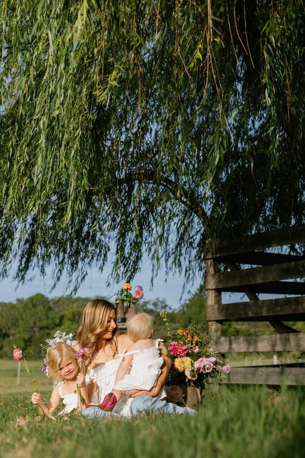 A woman with two young girls sitting on grass under a large tree with hanging branches, surrounded by colorful flowers and greenery.