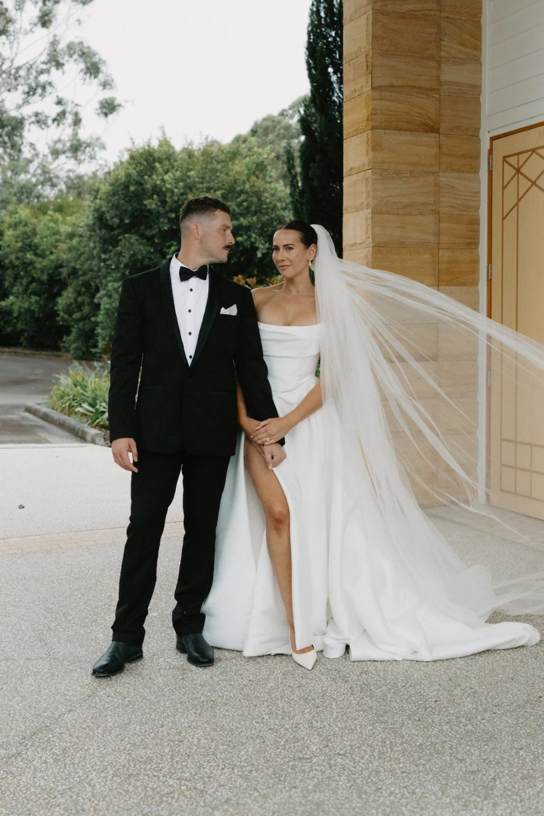 Bride and groom standing outside, bride in a white gown with a long train and veil, groom in a black tuxedo with a bow tie, holding hands, wedding dress revealing one leg, greenery and building in the background.