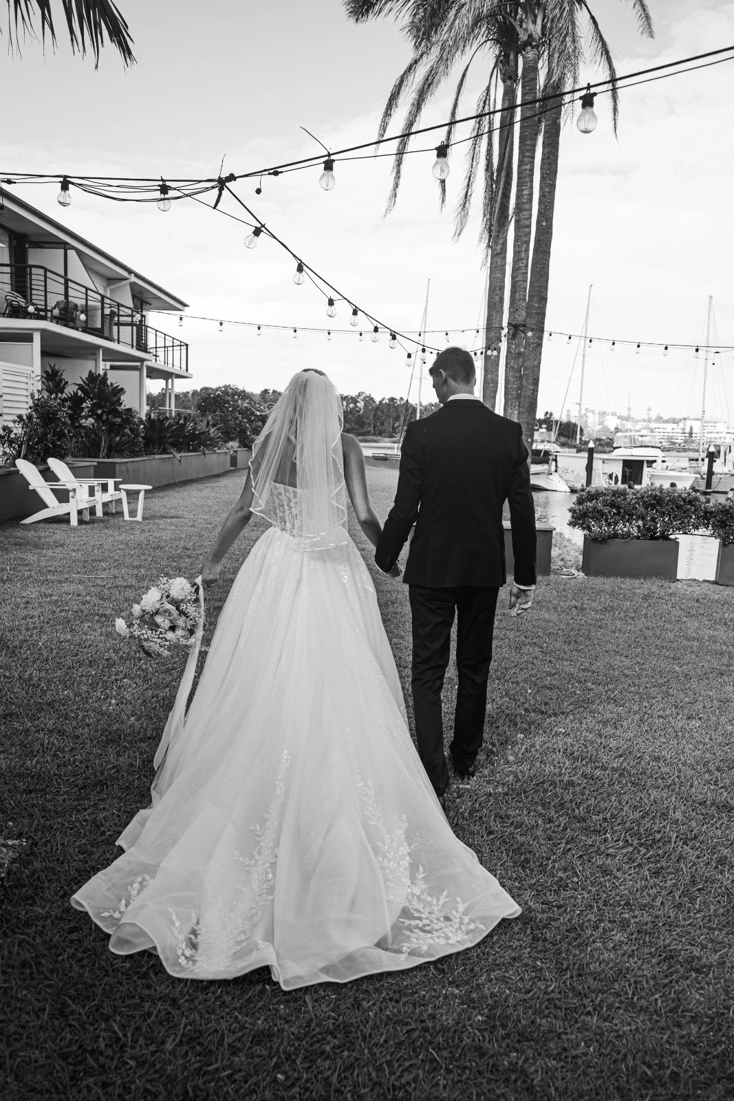 A bride and groom walking hand in hand along a waterfront lawn with palm trees, boats, and a string of outdoor lights in the background.