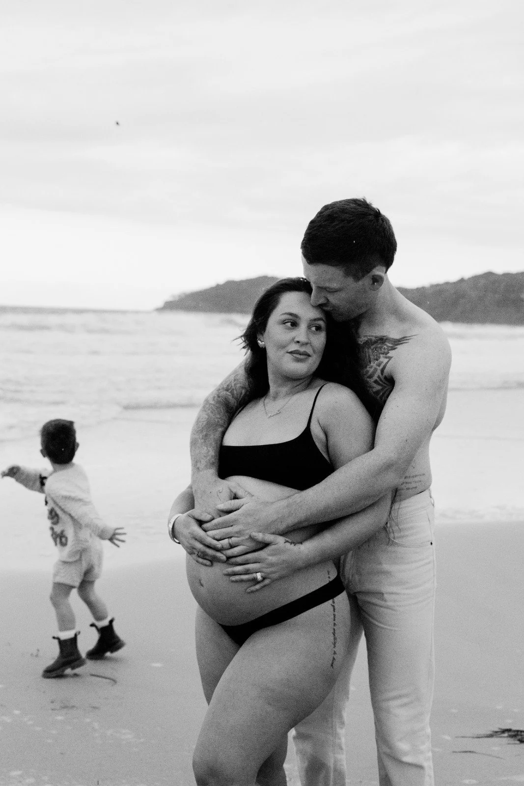 A pregnant woman and man embrace on a beach, with a child running in the background.