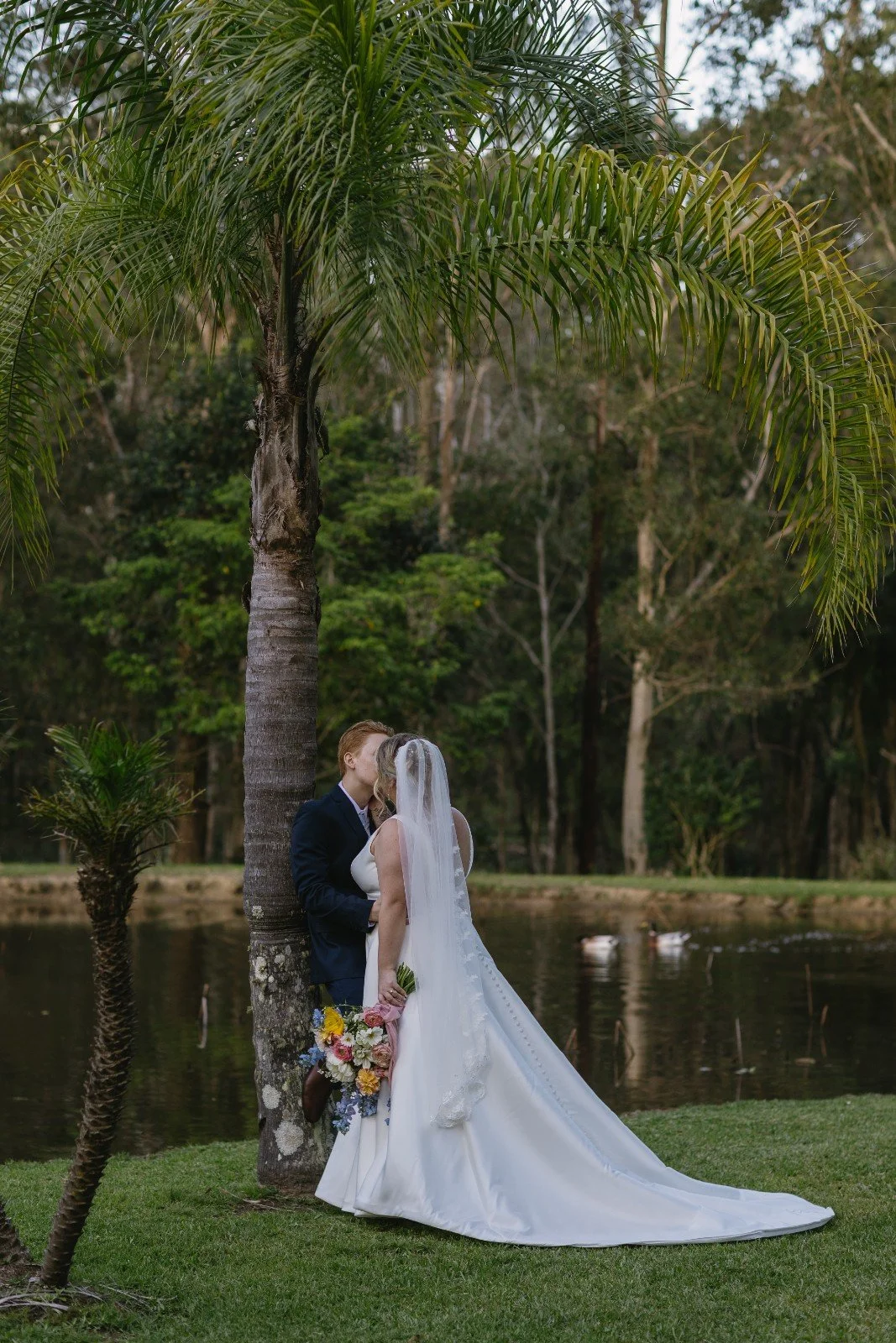 A bride and groom kissing behind a palm tree near a pond, with ducks swimming in the water and a forested background.