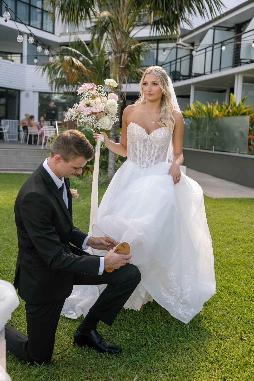 Bride in white wedding dress and groom in black suit on a grassy outdoor area with modern building and palm trees, groom kneeling and holding a shoe, bride holding bouquet.