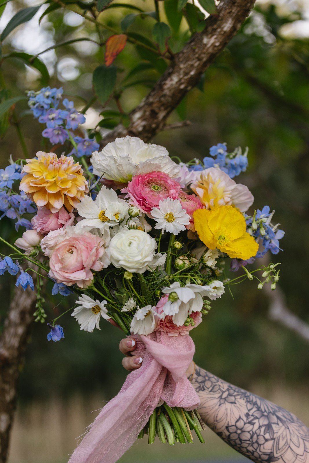 A hand holding a colorful bouquet of flowers tied with a pink ribbon, set outdoors with greenery in the background.
