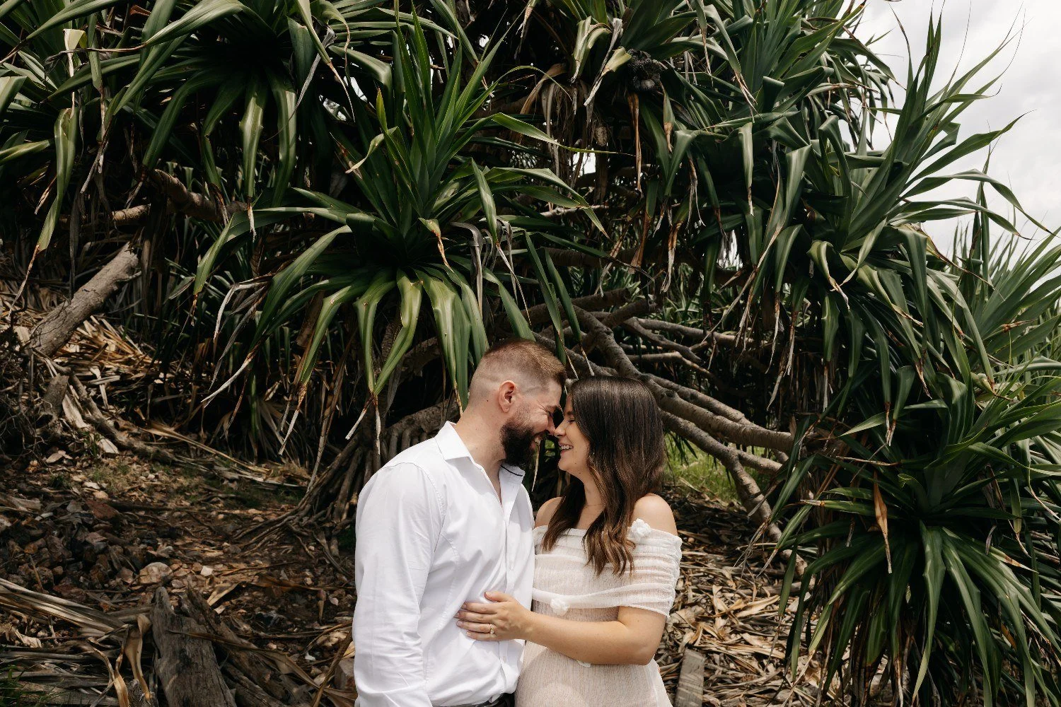 A happy couple with foreheads touching, smiling and holding hands in front of lush green tropical plants and roots.