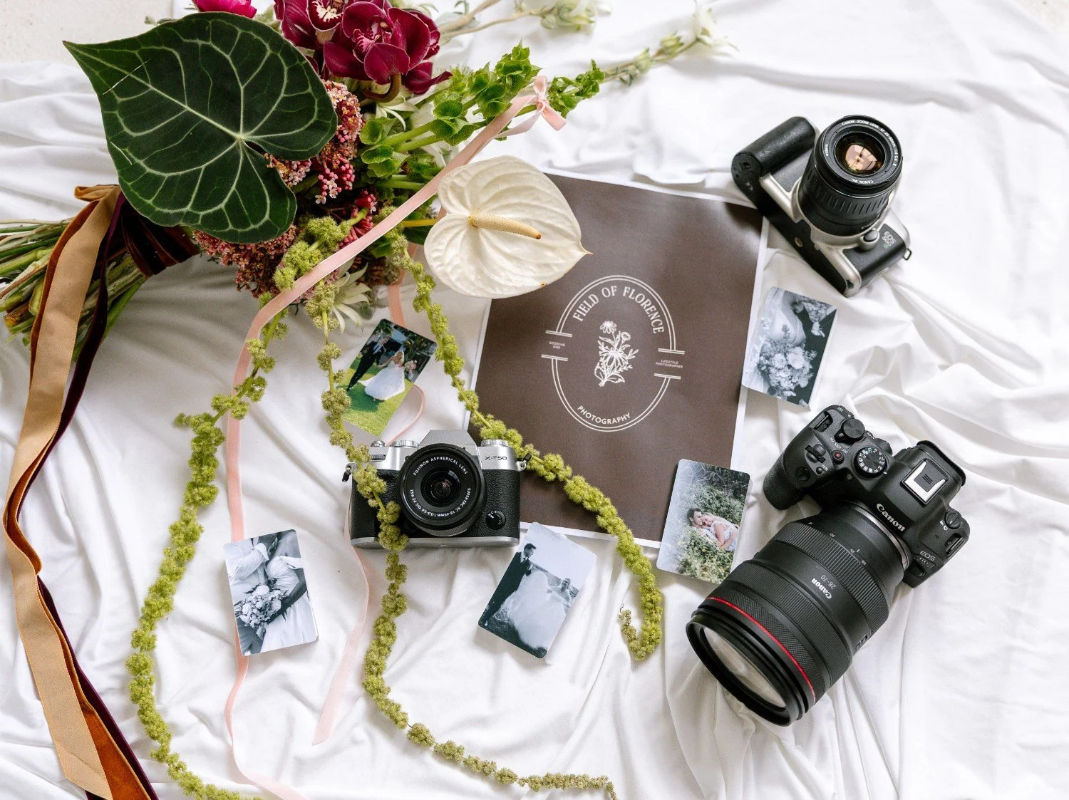 A flat lay of photography and floral items on a white fabric surface. It includes a bouquet of various flowers, two cameras, a book titled 'Field of Florence Photography,' printed photographs, and a vintage-style camera lens.