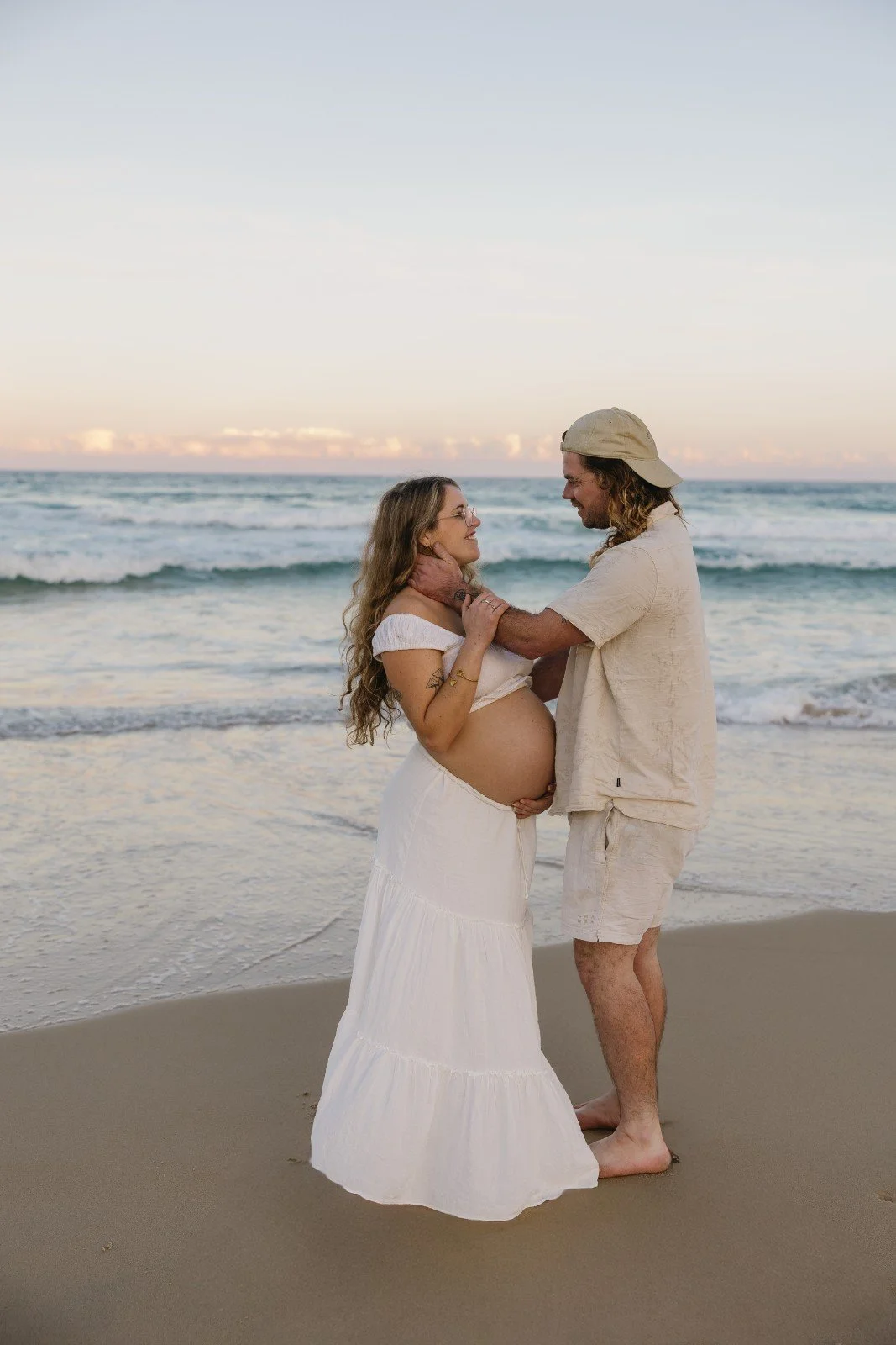 A pregnant woman and a man stand on the beach, smiling at each other. The woman is wearing a white off-shoulder dress, and the man is dressed in light-colored shorts and a shirt, with a backwards cap. The ocean and sky are visible in the background.