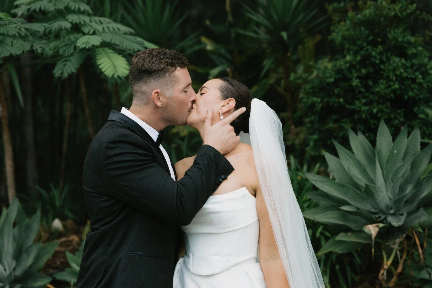 A newlywed couple kissing outdoors, surrounded by lush green plants and foliage.
