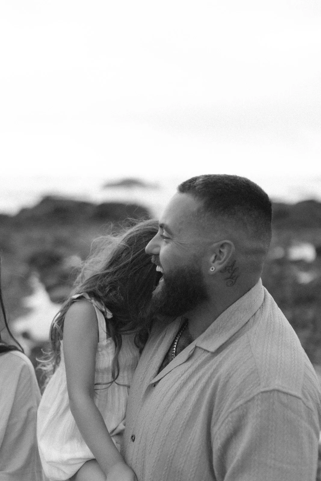 A black and white photo of a man and woman smiling and hugging outdoors, with the man facing right and the woman hugging him, her face hidden. Rocky terrain is visible in the background.
