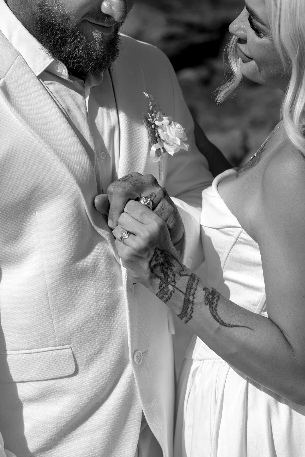 A close-up black and white photo of a couple holding hands during a wedding, with the groom in a light-colored suit with a boutonniere and the bride wearing a strapless dress with visible tattoos on her arm.