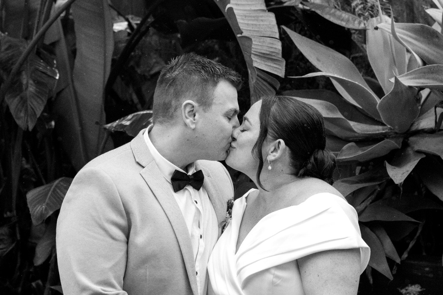 A black and white photo of a man and woman kissing in front of large leafy plants, dressed in formal attire, with the woman wearing pearl earrings.