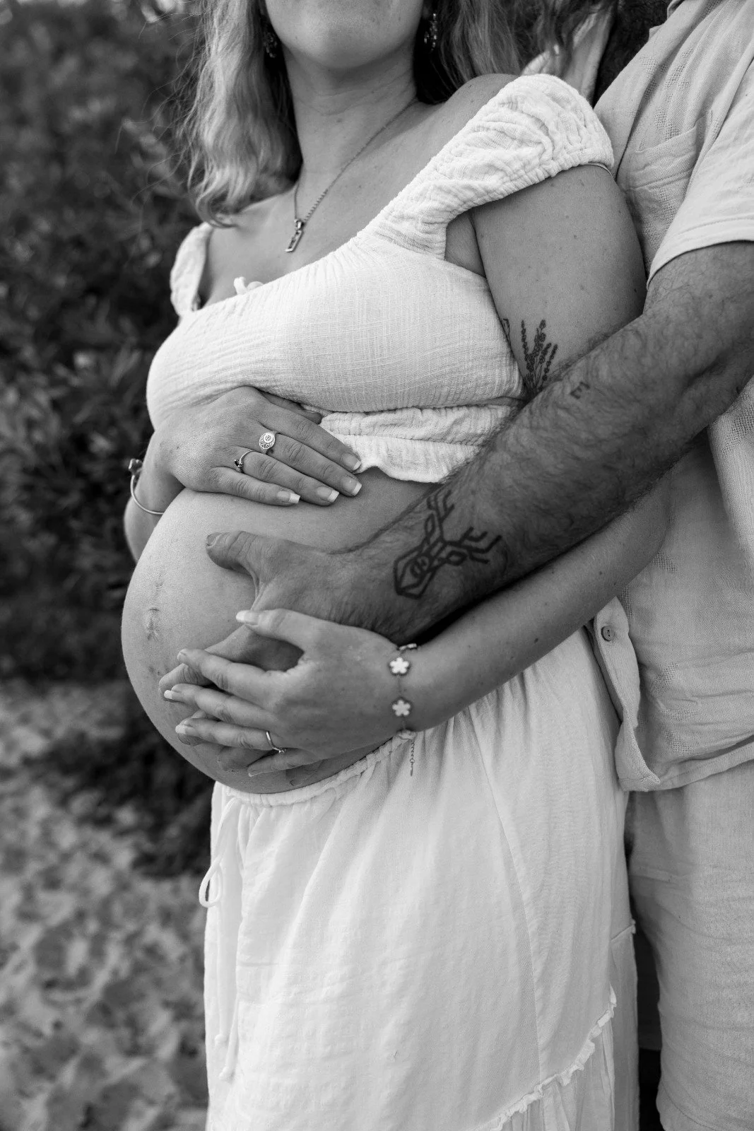 A pregnant woman and a man embracing, with their hands on her belly. Black and white photo, outdoors.