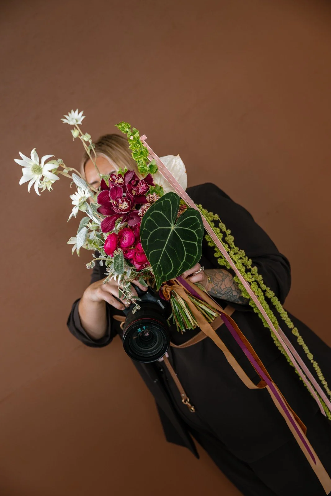 Person holding a colorful flower bouquet and a camera, standing against a brown background.