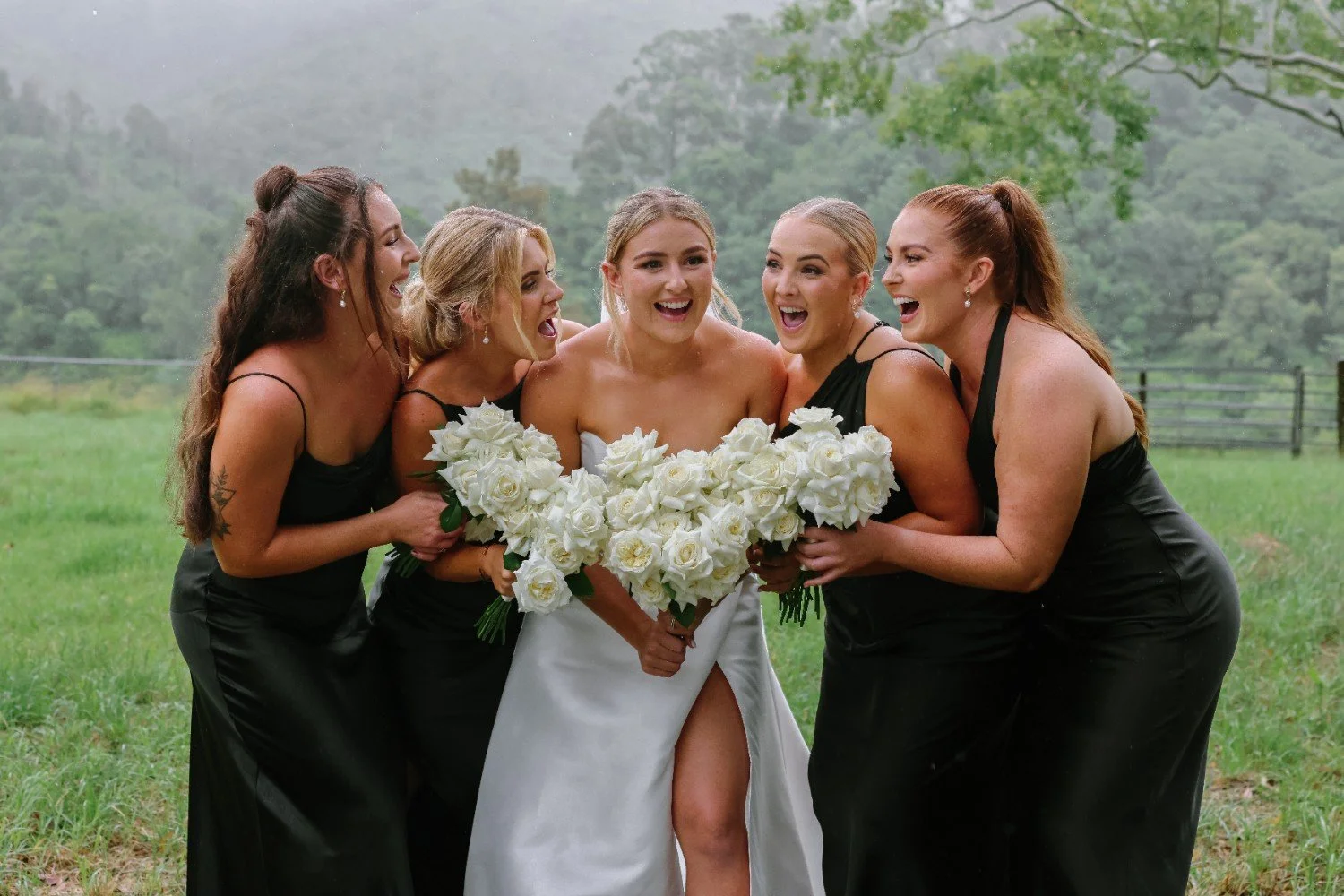 A bride in a white wedding dress holding a large bouquet of white roses, surrounded by five bridesmaids in black dresses, all smiling and laughing outdoors on a grassy field with trees in the background.