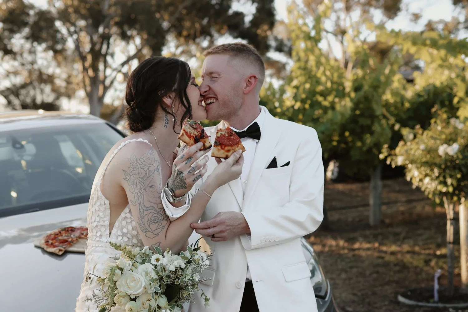 A newlywed couple with tattoos on the bride's arm, sharing a moment with pizza slices in an outdoor setting during sunset, standing next to a white car and surrounded by trees.