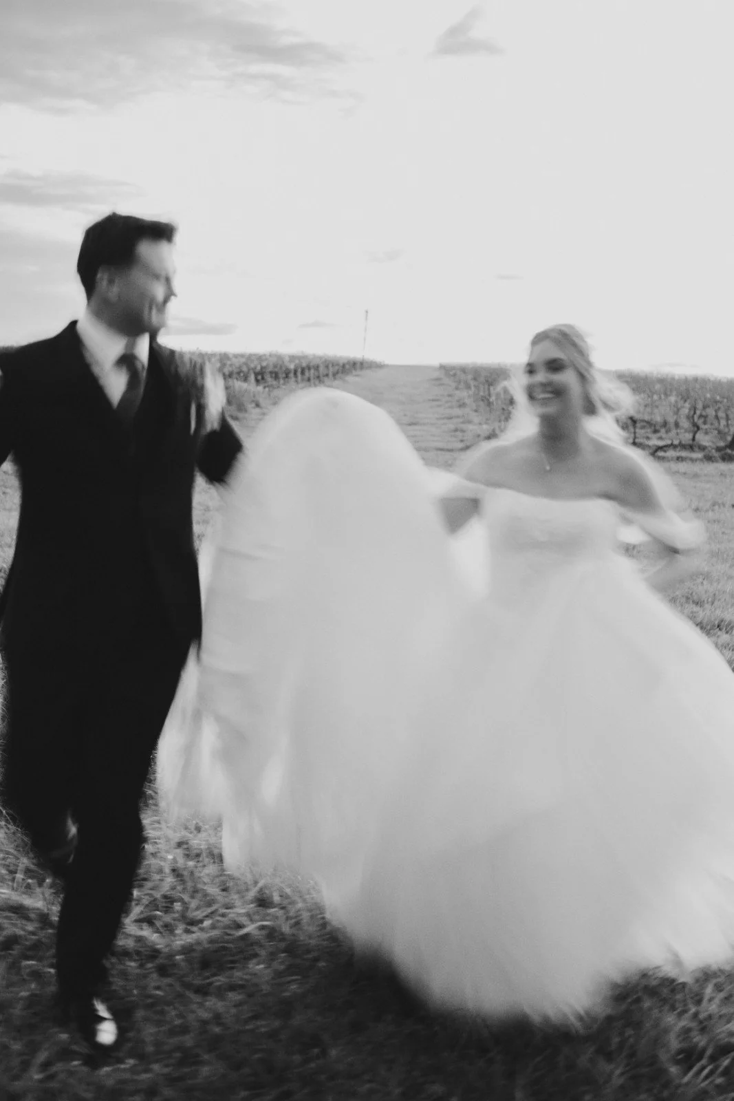 A black and white photo of a man and woman in a field, smiling and walking together. The woman is wearing a wedding dress, and the man is dressed in a suit.