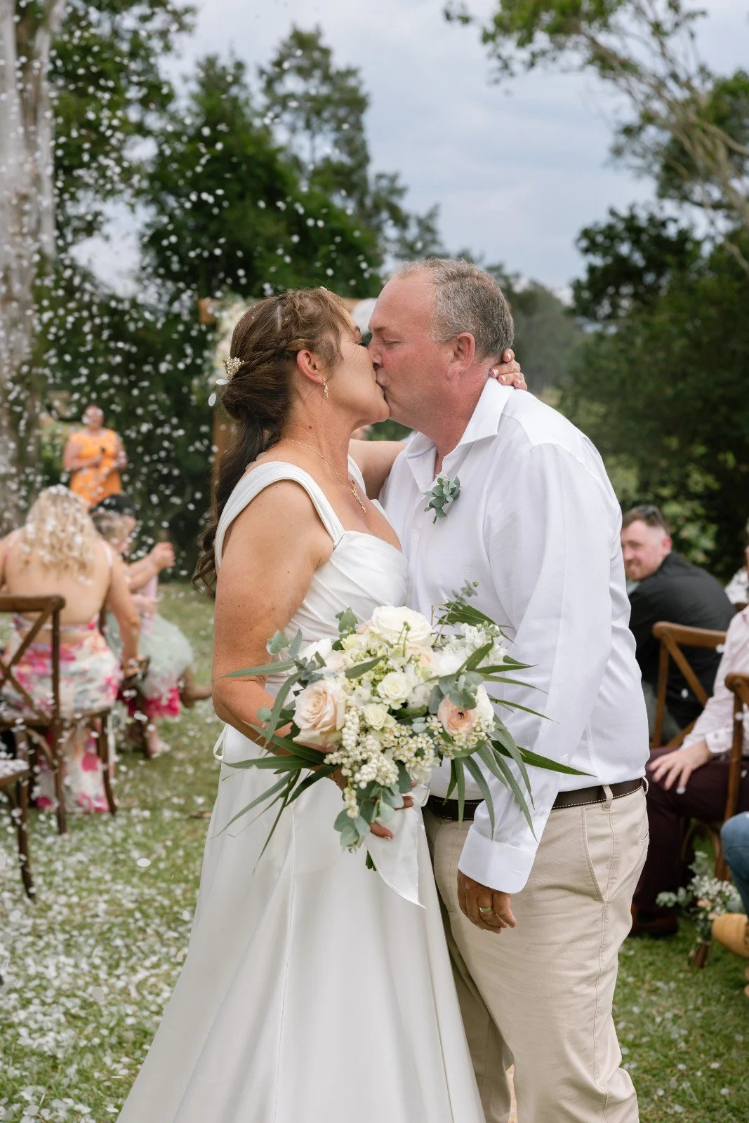 A couple in wedding attire sharing a kiss at an outdoor wedding, with guests and trees in the background.