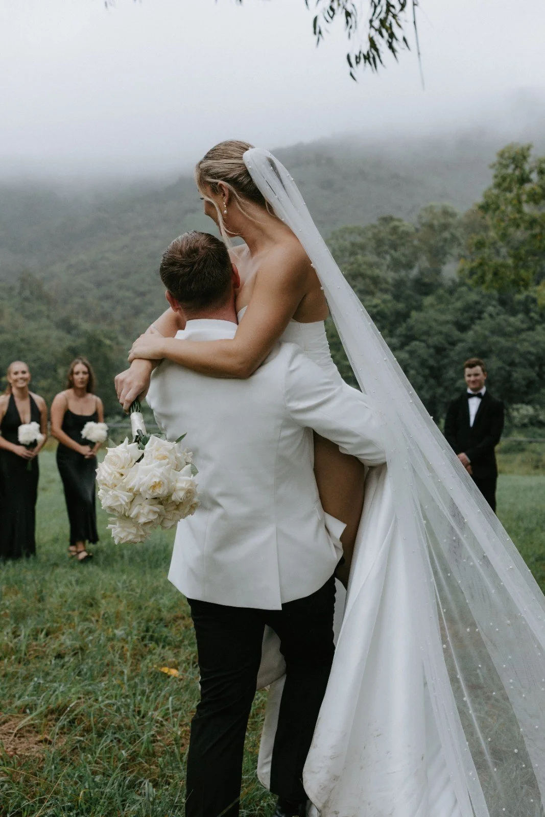 A groom holding a bride with a veil, on a grassy outdoor wedding ceremony, with bridesmaids and groomsmen in the background, surrounded by green mountains and cloudy sky.