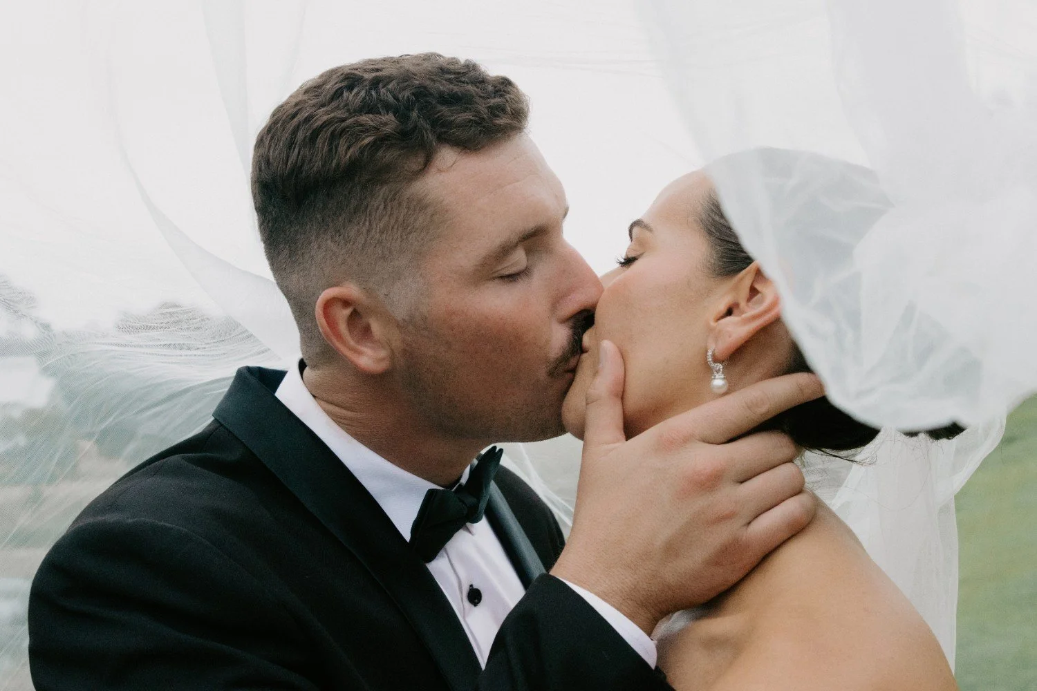 A bride and groom sharing a kiss outside, with the groom in a black tuxedo and the bride wearing a white dress and pearl earrings. The bride's veil is lifted by the wind.
