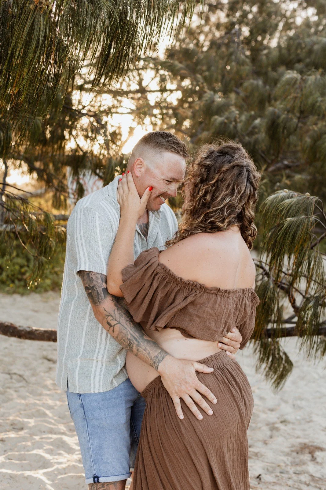 A man and a woman share an intimate moment on a sandy outdoor setting with trees. The man has short hair, tattoos, and is wearing a short-sleeved striped shirt and shorts. The woman has curly hair, is wearing an off-the-shoulder brown dress, and is t