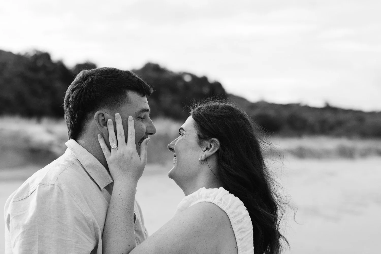 A couple sharing a joyful moment on the beach, with the woman touching the man's face and both smiling, in black and white.