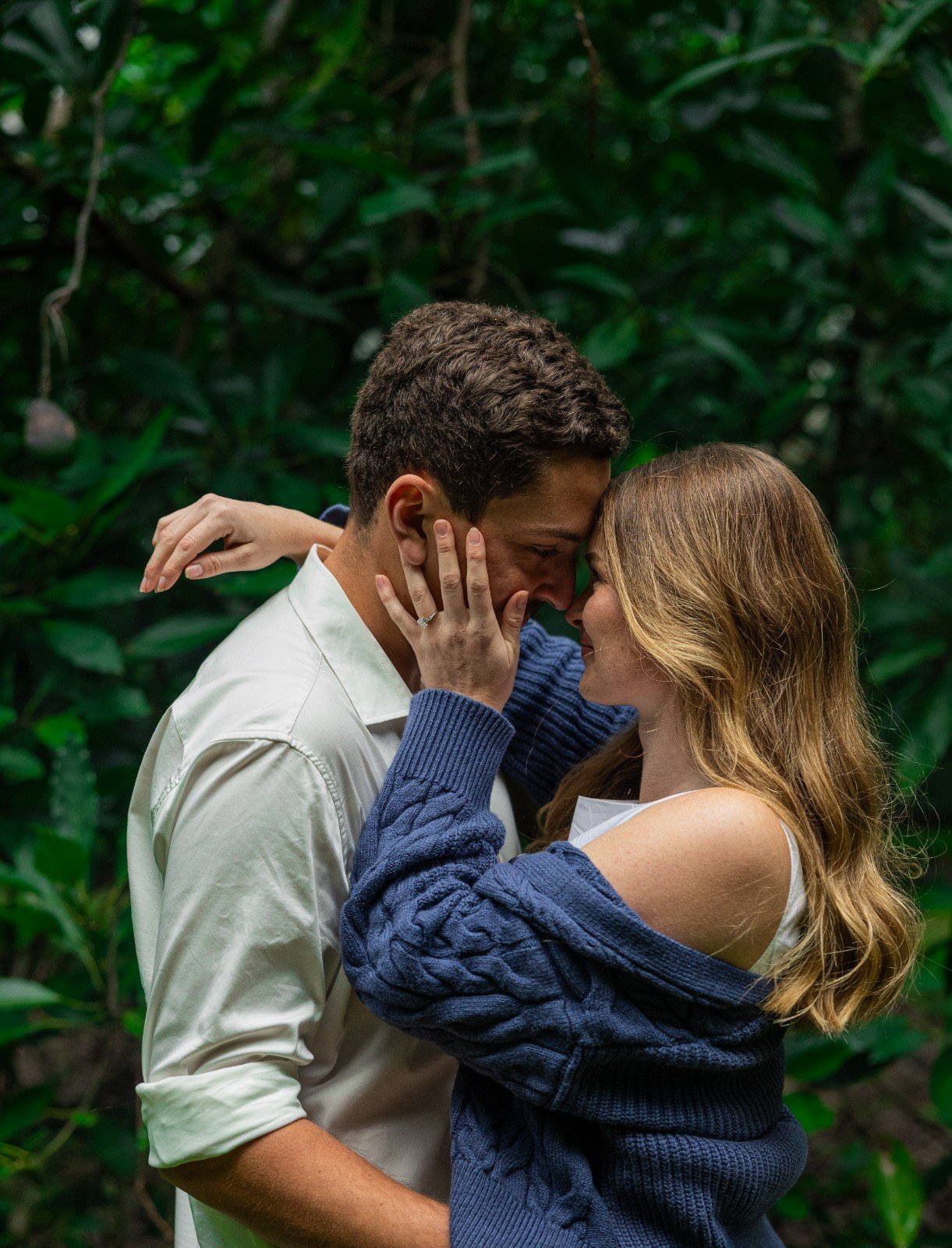 A romantic couple, a man and a woman, standing close in a lush green garden, touching foreheads and holding each other's faces gently.
