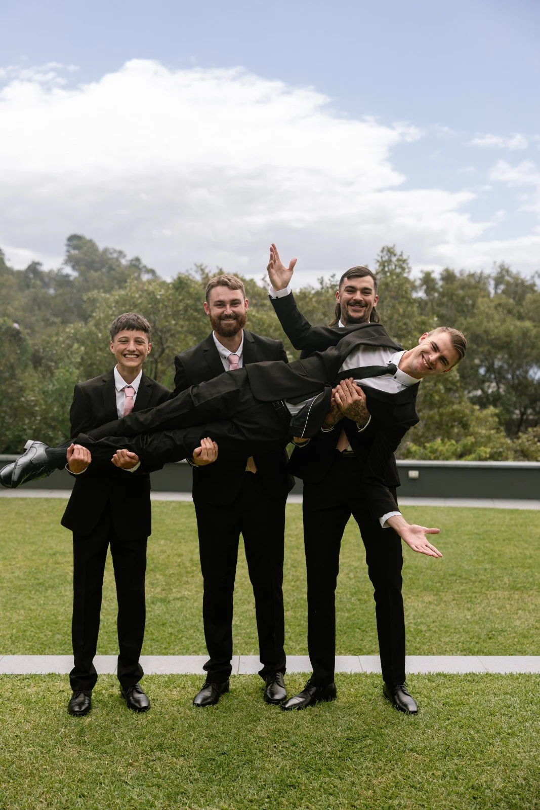 Four men dressed in suits, three smiling and holding up a fourth man horizontally in the air outdoors on a grassy field with trees and cloudy sky in the background.