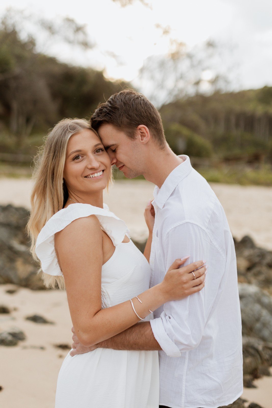 A happy couple embracing on a beach with trees and rocks in the background.