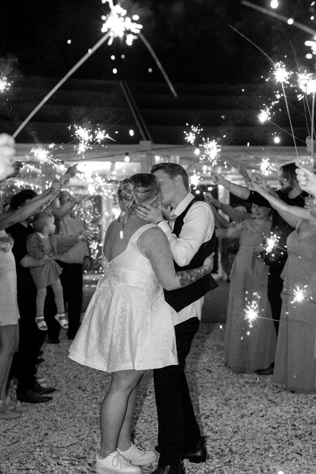 A couple sharing a kiss at a wedding reception, surrounded by guests holding sparklers.