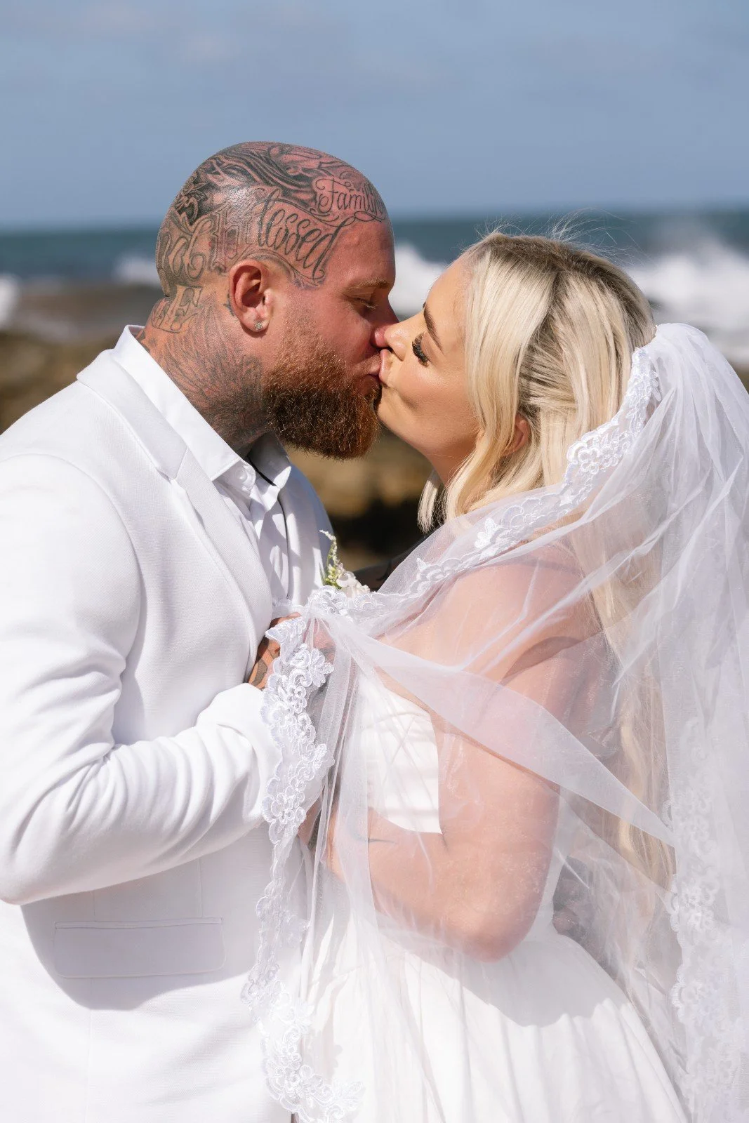 A newlywed couple is kissing on the beach with ocean waves in the background.