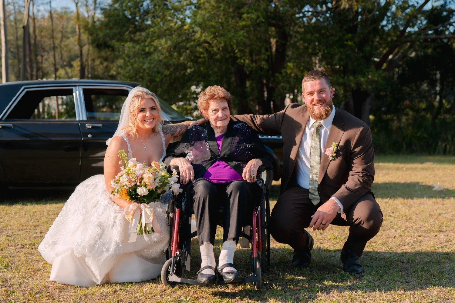 Bride, groom, and elderly woman in a wheelchair smiling outdoors during a wedding celebration with a vintage black car in the background.