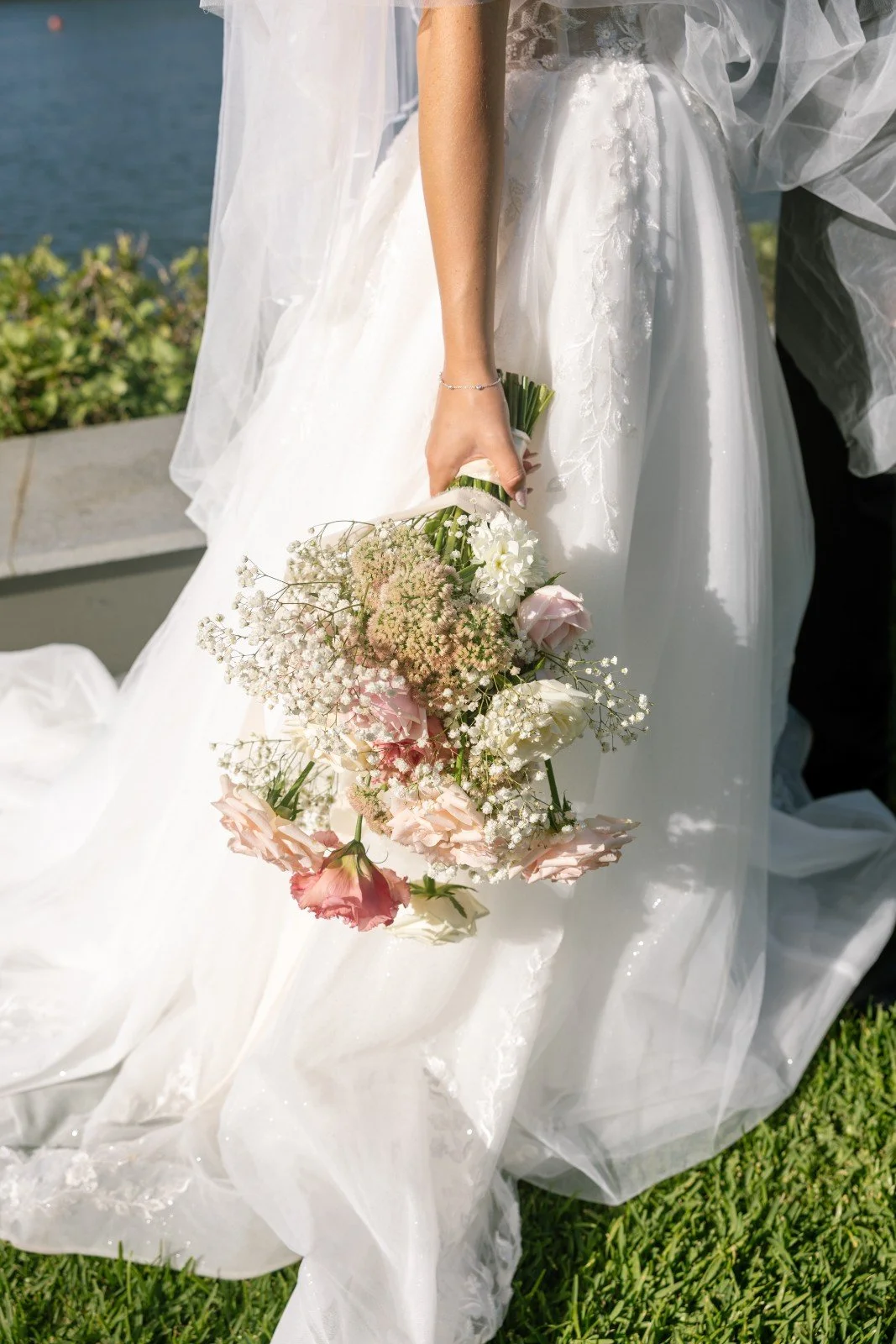 Close-up of a bride holding a bouquet of white and pink flowers, wearing a white wedding gown and veil, outdoors near water.